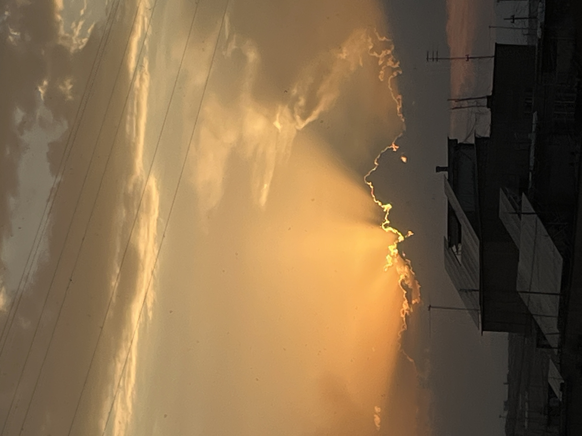 The image depicts a dramatic sunset over a cityscape, possibly in Yerevan, Armenia. The sky is ablaze with warm orange and yellow hues, as the sun's rays fan out from behind a large, dark cloud formation. The sunlight illuminates the edges of the clouds, creating a vibrant contrast against the darkening sky.
In the foreground, silhouetted against the bright sky, are the rooftops of several buildings. These structures appear to be utilitarian, possibly residential or industrial, with flat roofs and some visible ventilation or antenna systems. The overall impression is one of urban sprawl meeting a powerful natural phenomenon. The presence of power lines stretching across the upper portion of the frame further emphasizes the urban setting. The image has a slightly grainy texture, suggesting it was taken with a mobile device or in low light conditions.
