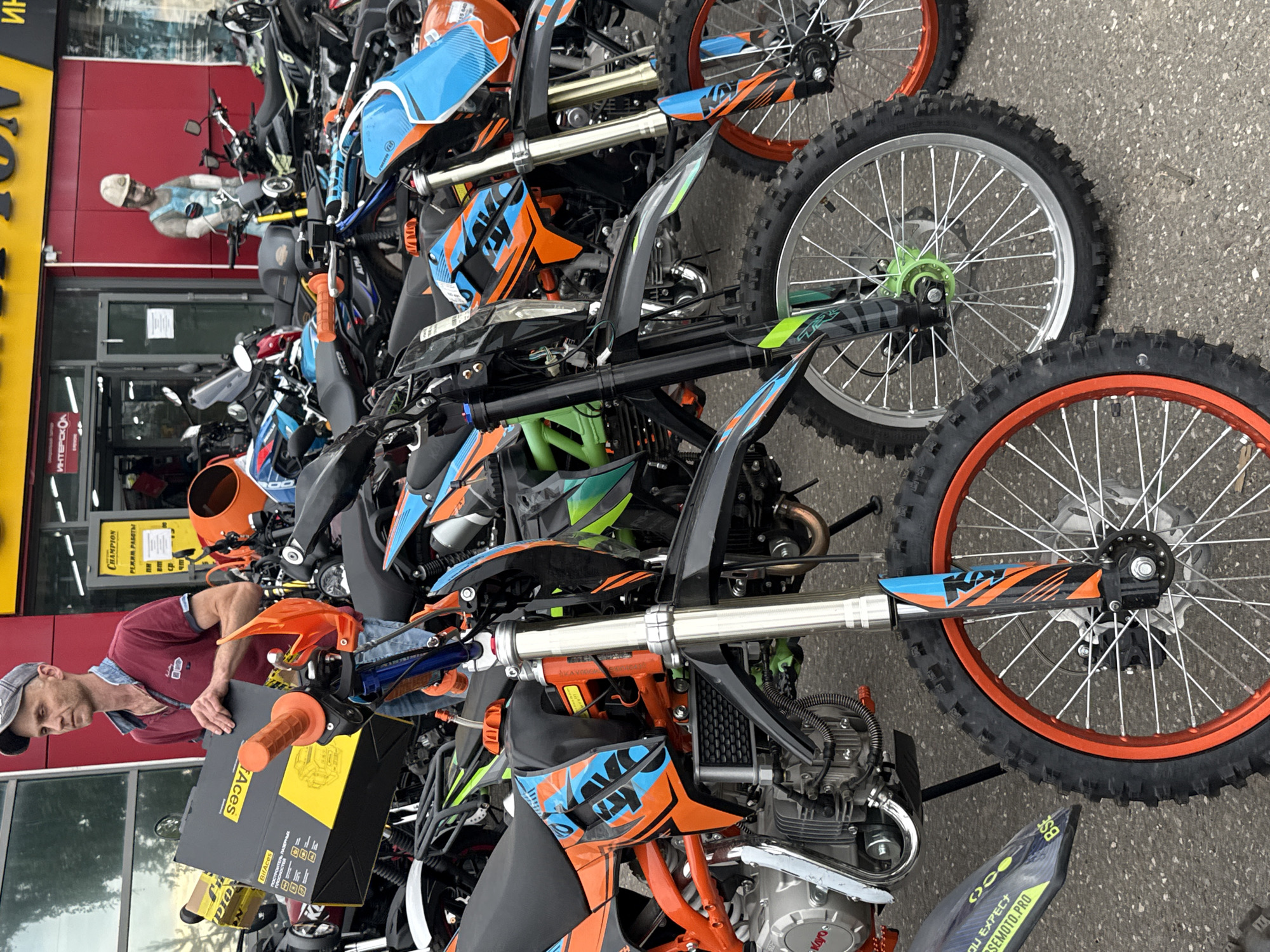 The image captures a street-level view in front of a store in Votkinsk, Russia. A man wearing a maroon polo shirt and a grey baseball cap is holding a large black and yellow box labeled "BitAces" and "Enduro." He is standing amongst a collection of dirt bikes, some of which are orange and blue. The store behind him has a red facade and a large yellow sign with Russian text that appears to advertise a business. A mannequin dressed as a construction worker stands outside the store. The ground is asphalt, and the overall scene suggests a retail environment focused on vehicles or equipment.