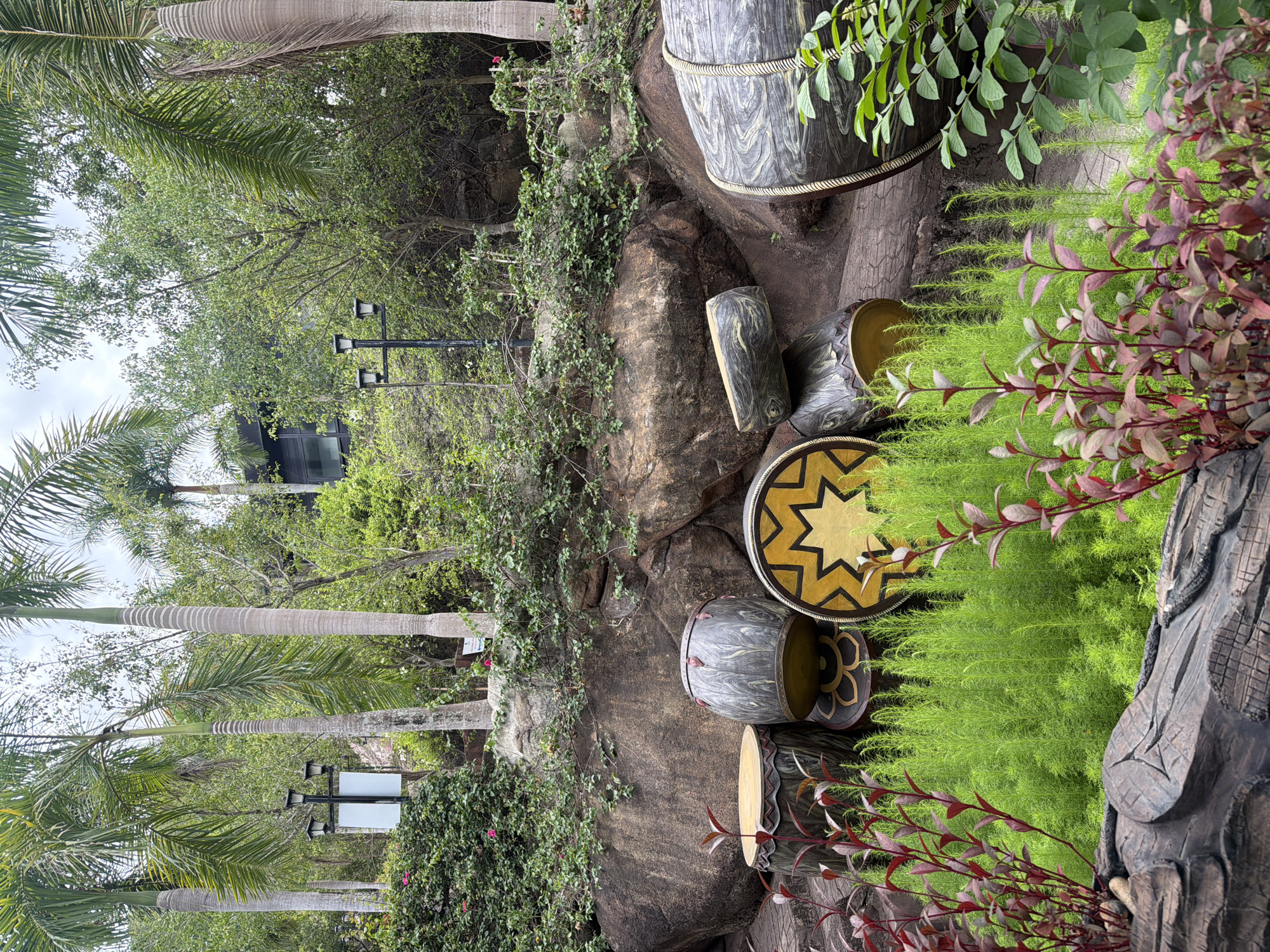 The image depicts an outdoor scene with several large, decorative drums resembling those used in traditional ceremonies. These drums are arranged on a rocky slope covered with lush greenery. Palm trees are visible in the background, along with some architectural elements, including what appears to be a building with dark-tinted windows and a lamppost. In the foreground, there is dense green foliage and some plants with reddish-purple leaves. The overall setting suggests a park or garden area. This scene is located in the VinWonders Nha Trang amusement park on Hon Tre Island, Nha Trang, Vietnam.