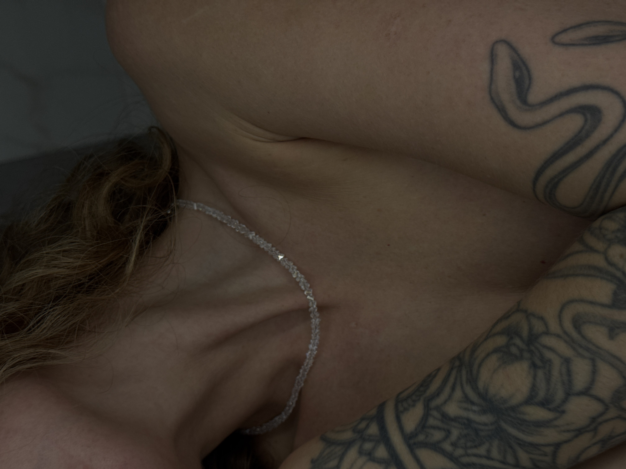 The image displays a close-up, partial view of an individual's upper torso, neck, and a portion of their head. The person has fair skin and wavy, light brown or blonde hair, visible on the right side of the frame. They are wearing a delicate choker-style necklace made of small, clear or translucent faceted beads, with a single, small metallic, geometric bead accenting the chain near the front.
Extensive tattoos are visible on both arms. On the subject's right arm (viewer's left), there is a detailed sleeve tattoo featuring intricate floral or mandala-like patterns rendered in dark ink. On the subject's left arm (viewer's right), a more minimalist, outline-style tattoo of a snake is present, with its head directed upwards. The person's arms appear to be crossed or held in a relaxed position across their body.
The lighting is soft and somewhat dim, casting subtle shadows that highlight contours of the skin and hair. The background is uniformly dark, indistinct, and out of focus, suggesting an indoor setting with a plain wall or fabric behind the subject. There is no visual information within the image to determine its geographic location, including any indication of the city of Chaikovsky, Russia.
