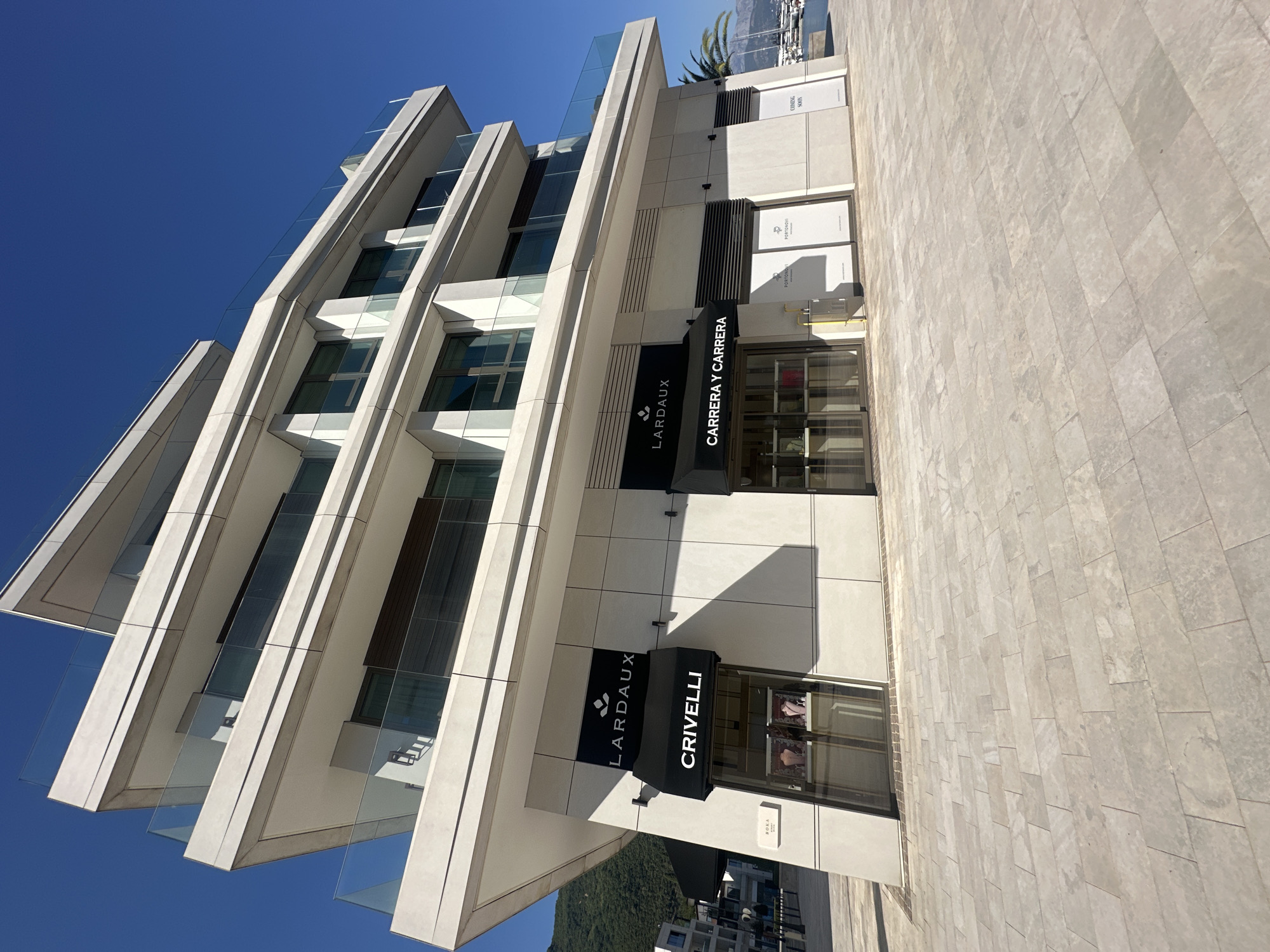 A low-angle, exterior, full shot captures the modern, multi-story facade of a building on a sunny day in Kumbor, Montenegro. The building features a contemporary architectural design with sharp angles, cantilevered balconies with glass railings, and large windows. On the ground level, there are several commercial establishments. Two prominent awnings are visible: one displaying "LARD" (partially obscured) and "CRIVELLI" in white lettering on a black background, and another with "LARD" (partially obscured) and "CARRERA Y CARRERA" in white lettering on a black background. Below the "CRIVELLI" awning, a shop window displays jewelry. Adjacent to this, two windows are covered with white signs featuring the "PORTONOVI" logo. Further to the right, another window has a "COMING SOON" sign. The ground in front of the building is paved with light-colored rectangular stones. The background shows a clear blue sky, a hint of a mountainside to the left, and a harbor with sailboats to the right. There are no people visible in the frame.