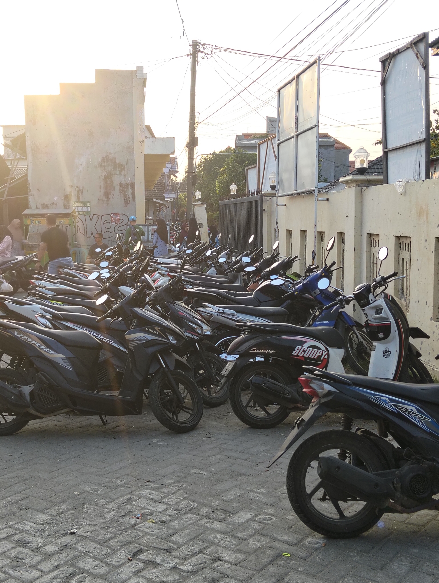 A low-angle, close-up shot captures a densely packed parking lot filled with parked motorcycles. The motorcycles, predominantly black and grey, are lined up in multiple rows on a paved surface. Several are visible with "Honda" branding on their front panels. Some are partially obscured by others. In the background, a beige-colored building with a rough, textured facade is seen, catching the warm, golden light of what appears to be late afternoon or early morning sun, which casts lens flares across the image. Several individuals, including women wearing headscarves and men, are casually standing or walking among the motorcycles. One man in a black shirt and shorts is seated. The scene is set against a backdrop of utility poles and overhead wires, common in urban Indonesian settings. The context suggests a busy area, possibly near a marketplace or event venue in Banjarwati, Indonesia, indicated by the visible text "SEGO MUDUK ENDANG 7.000" on the building, which translates to "Rice dish of Endang 7,000", a common local food stall sign. The overall atmosphere is one of everyday life in a bustling Indonesian town.