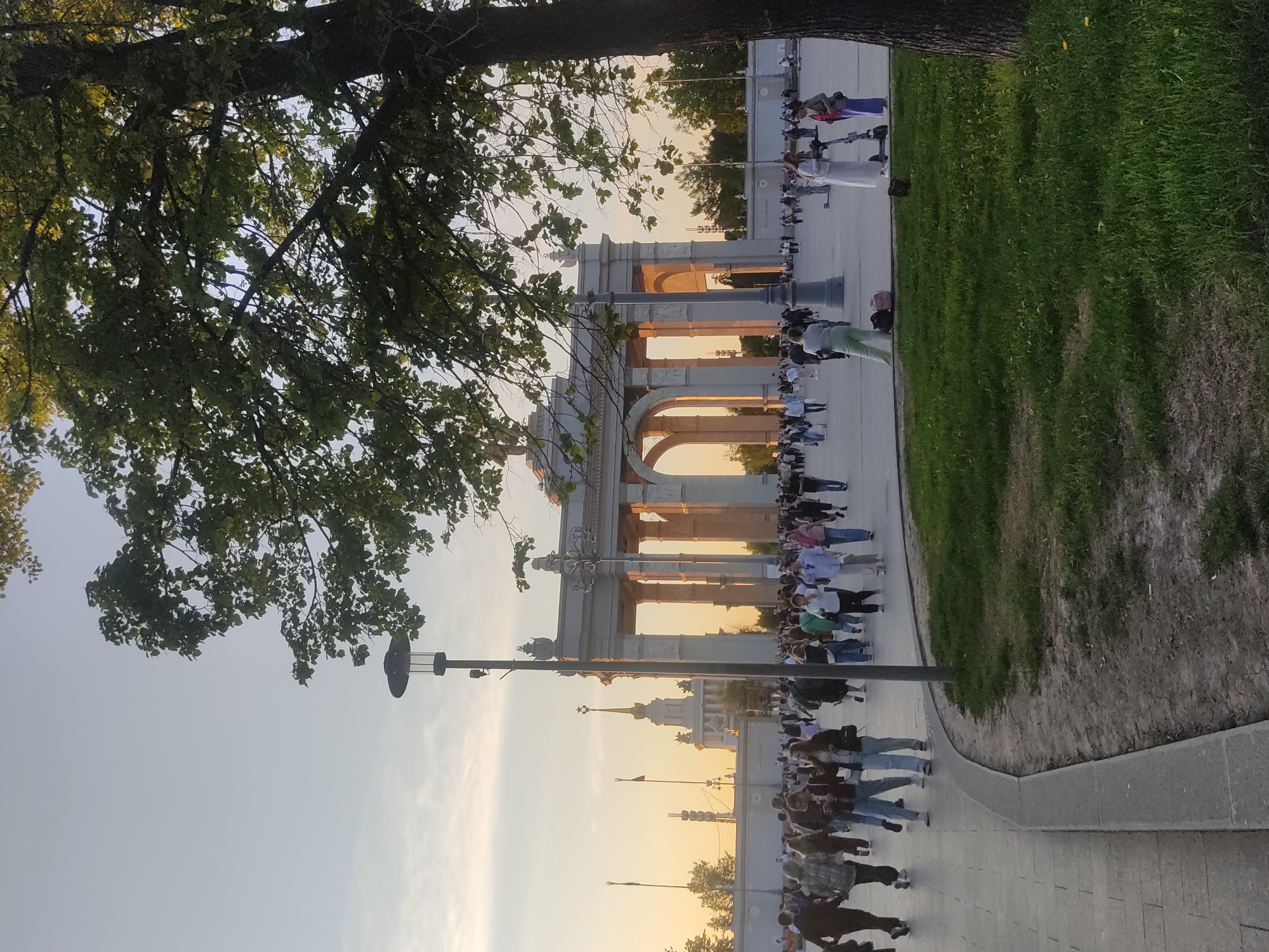 A long line of people walks past the Triumphal Arch in Moscow, Russia. The arch is composed of a series of arches with golden light filtering through. A tree is in the foreground on the left. Grass is to the right of the sidewalk, and buildings are visible in the distance.