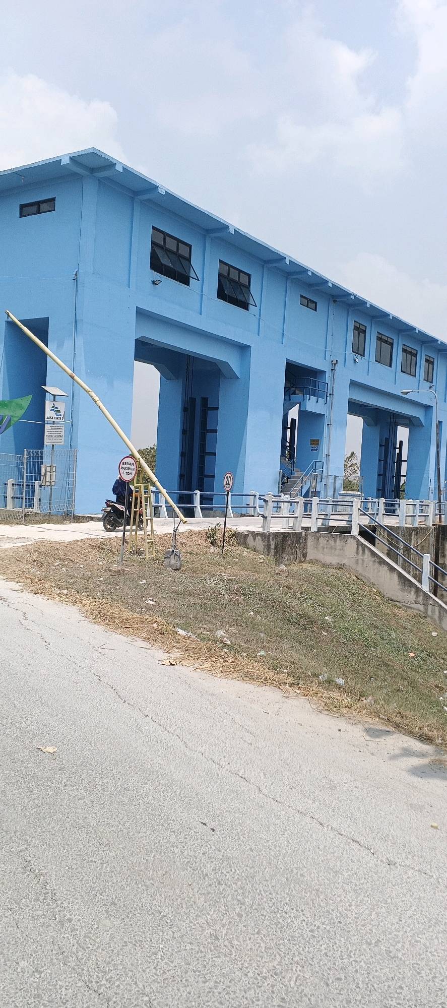 This is a landscape orientation, eye-level, medium shot image of the P.J.A. Tirta II dam in Parengan, Indonesia. The dam is a large, light blue concrete structure with several arched openings, some of which have visible vertical gates. The building has multiple rectangular windows, many of which are open. A yellow barrier arm is extended across a road leading to the dam. To the left of the barrier, a sign for "JASA TIRTA II" and "STASIUN P.J.A. TIRTA II" is visible. A red and white circular sign with "20" is posted on a pole near the barrier. A single motorcycle with a rider is parked to the left of the barrier. The ground in front of the dam is covered with dry grass and some litter. A paved road with some cracks is in the foreground, leading away from the dam. The sky is cloudy.