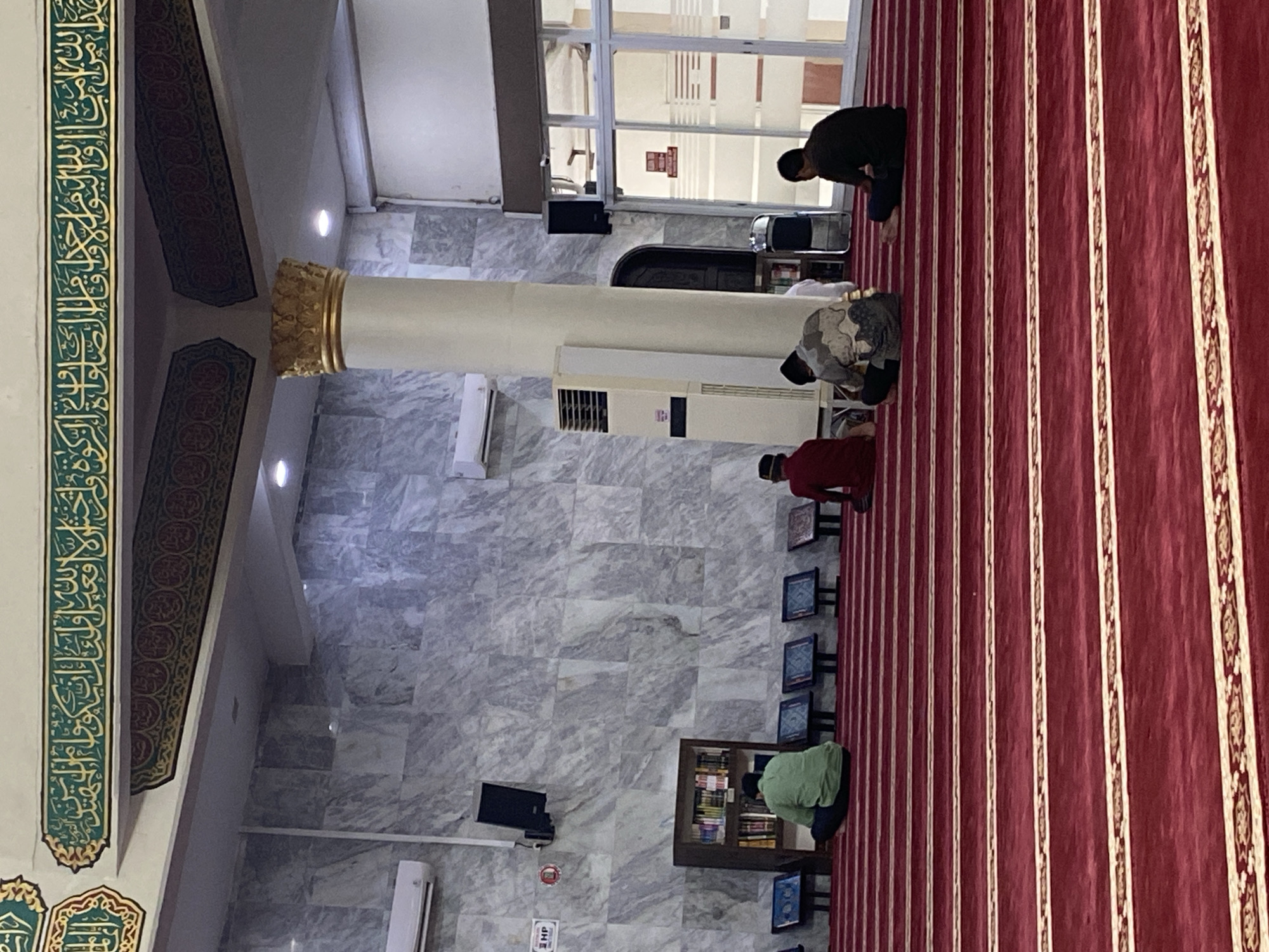 The image depicts the interior of a mosque, likely located in Lhokseumawe, Indonesia. The foreground is dominated by a richly carpeted floor with distinct red rows separated by decorative gold and cream stripes. In the middle ground, four individuals are seated on the carpet, facing away from the camera, engaged in prayer or contemplation. One person is wearing a green shirt, another a red shirt, a third a patterned shirt, and the fourth a dark shirt. To the left, a bookshelf filled with books stands against a marbled wall. Above, ornate calligraphy in green and gold adorns the upper wall and ceiling, a common feature in Islamic architecture. Air conditioning units are visible on the walls, and a large white column with a gold capital stands prominently. In the background, large glass doors lead to another area of the mosque. The overall atmosphere is one of quiet reverence and devotion.
