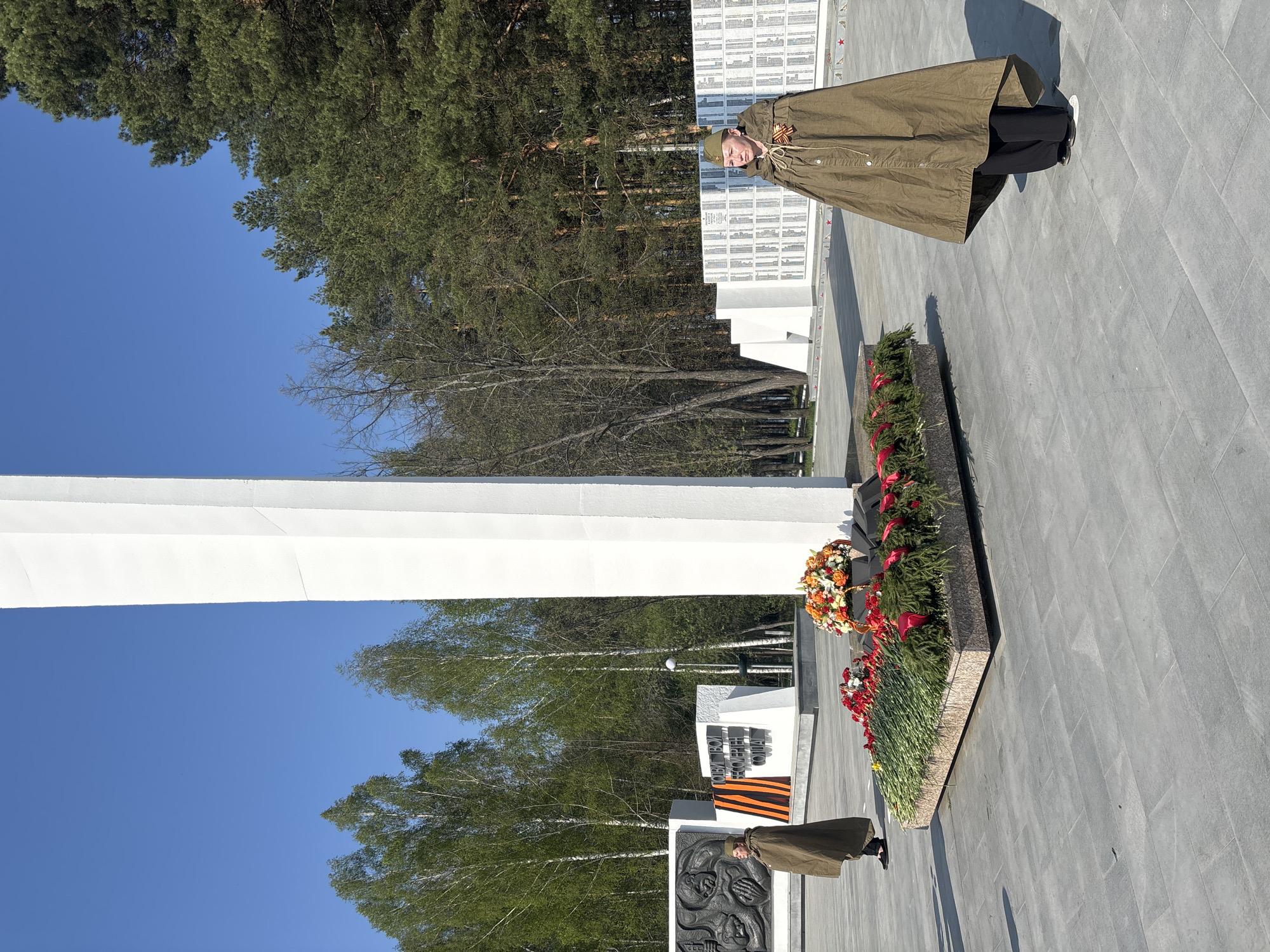 A person wearing a green military-style cape and cap stands in front of a World War II memorial in the city of Chaikovsky, Russia. The memorial includes a tall white obelisk, engraved stone monuments, a wall of names, wreaths, and flower arrangements. The sky is blue and trees surround the memorial site. The ground is paved with gray stones.