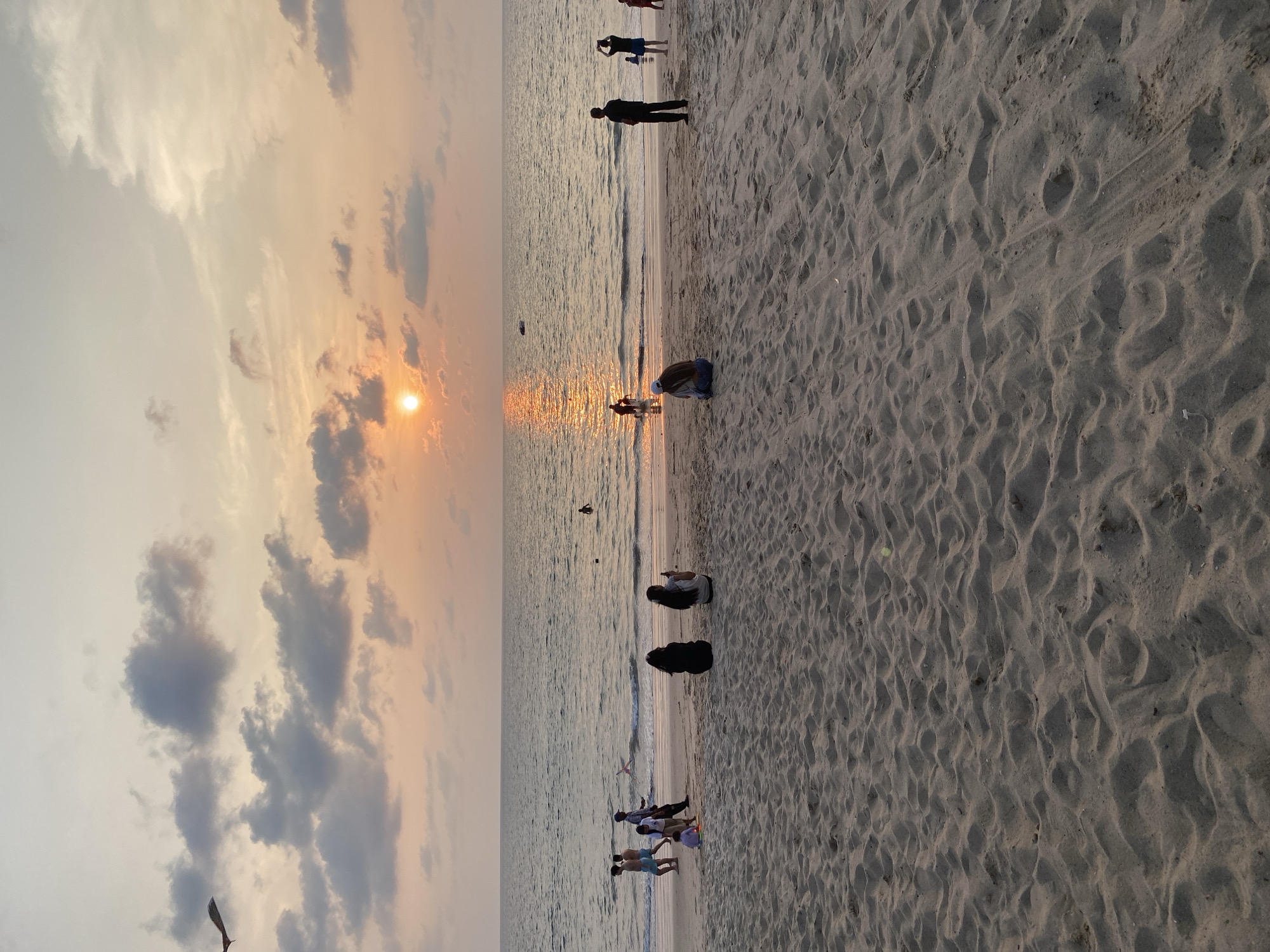 The image depicts a beach scene at sunset in Choeng Thale, Thailand. Several people are present, some standing along the shoreline and others walking or sitting on the sand. The sky is filled with clouds, and the sun is partially obscured, creating a warm glow over the water. The beach is sandy, with footprints and patterns visible on the surface.