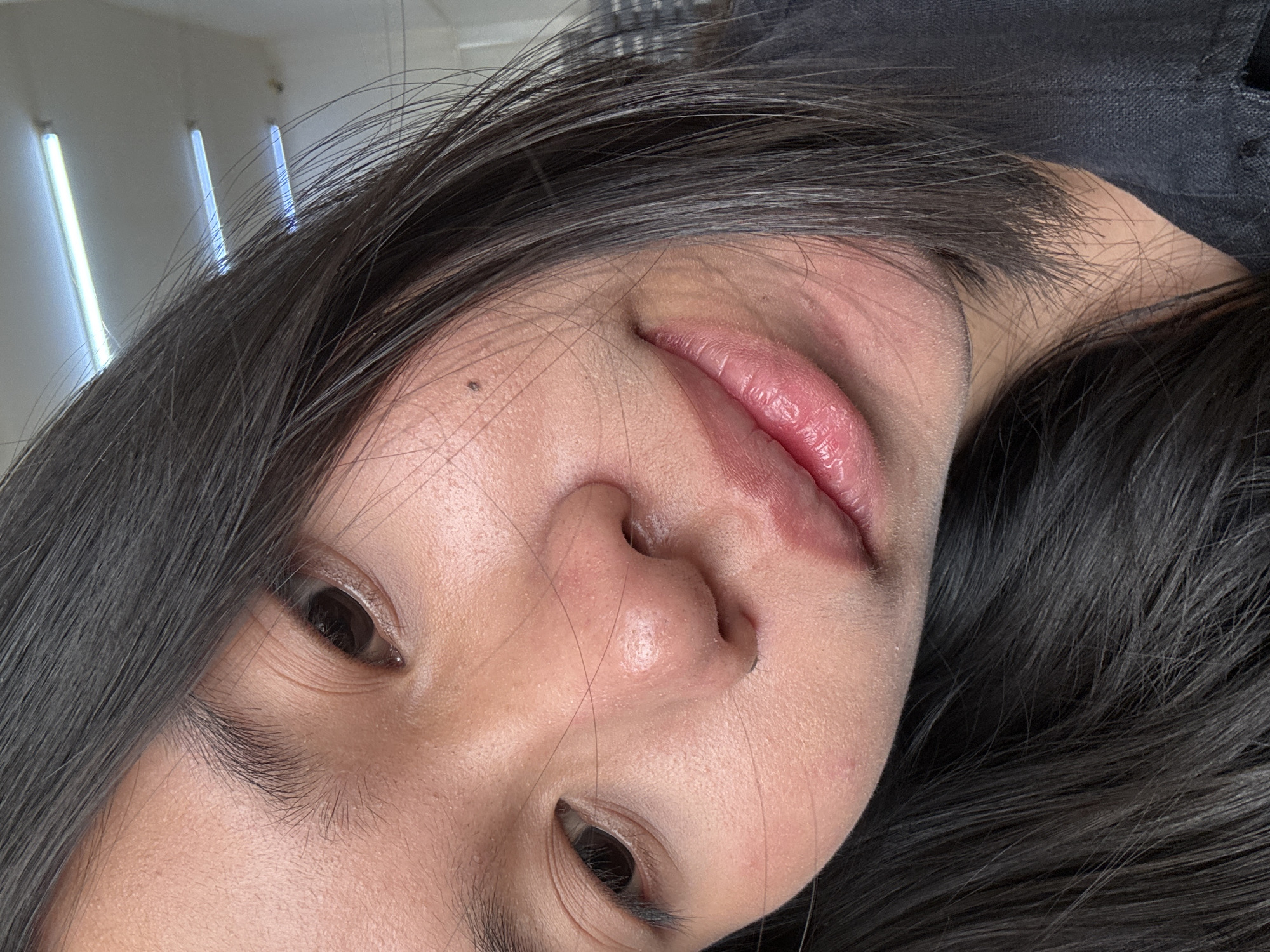 A close-up, head and shoulders shot captures a young East Asian woman in a casual, indoor setting. She has dark hair that frames her face, and her gaze is directed towards the camera. Her skin appears to have a light dusting of what might be powder or fine debris. The background is out of focus, but appears to be an indoor space with fluorescent lighting visible on the ceiling. A portion of a dark blue garment, possibly a shirt or jacket, is visible on her right side. The overall lighting suggests an ambient indoor environment. There is no indication of the specific location within East Jakarta, Indonesia.