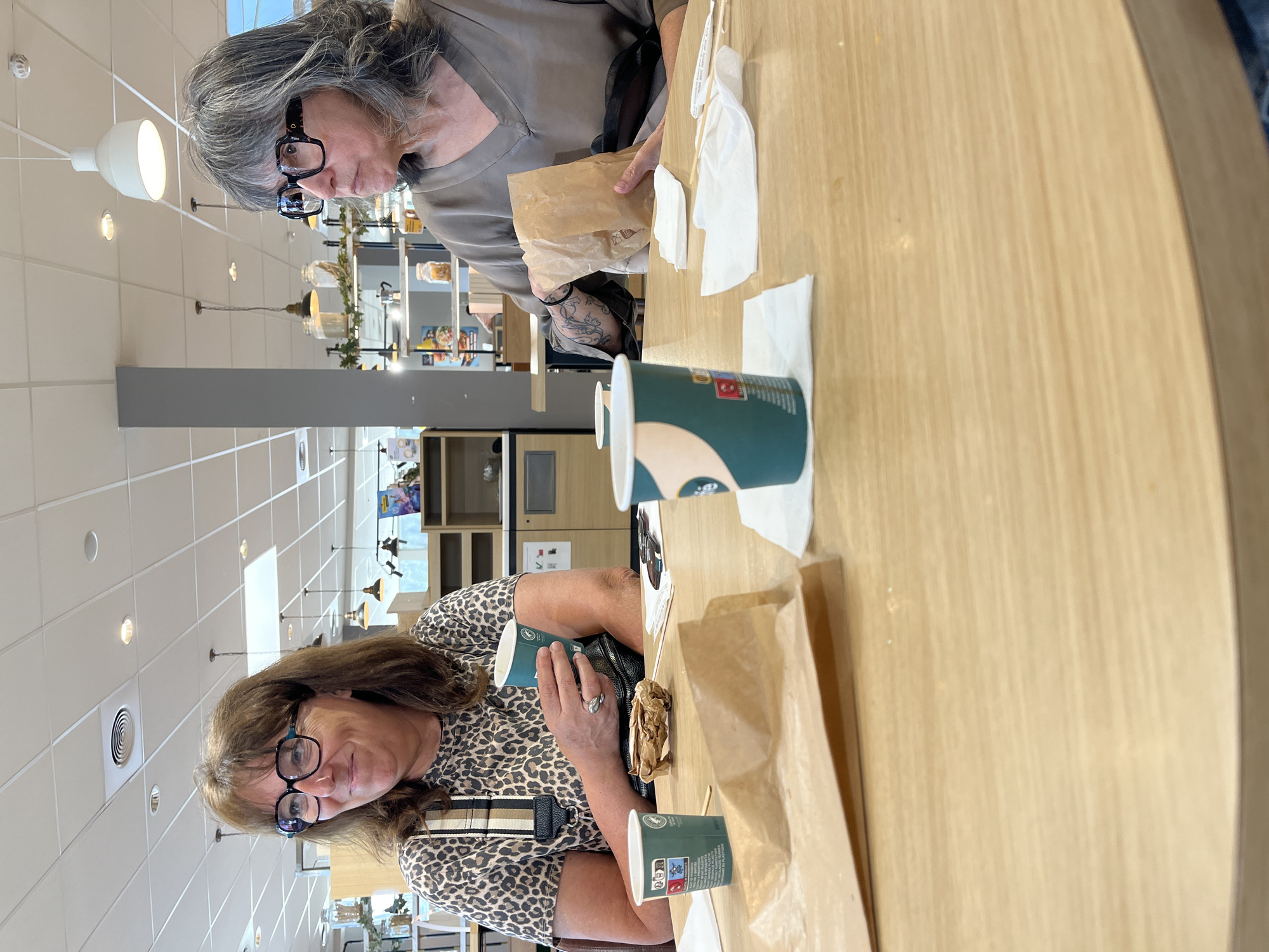 The image shows two women sitting at a light-colored wooden table in what appears to be a restaurant or cafe. The woman on the left wears a leopard-print top and glasses. She holds a green and white paper cup. Next to her is a crumpled paper bag. The woman on the right has gray hair and wears glasses and a light brown top. She holds a similar paper bag. There are other green and white paper cups and napkins on the table. The background includes a view of the restaurant interior with overhead lighting, shelves with items, and some greenery. The location is Rousset, France.