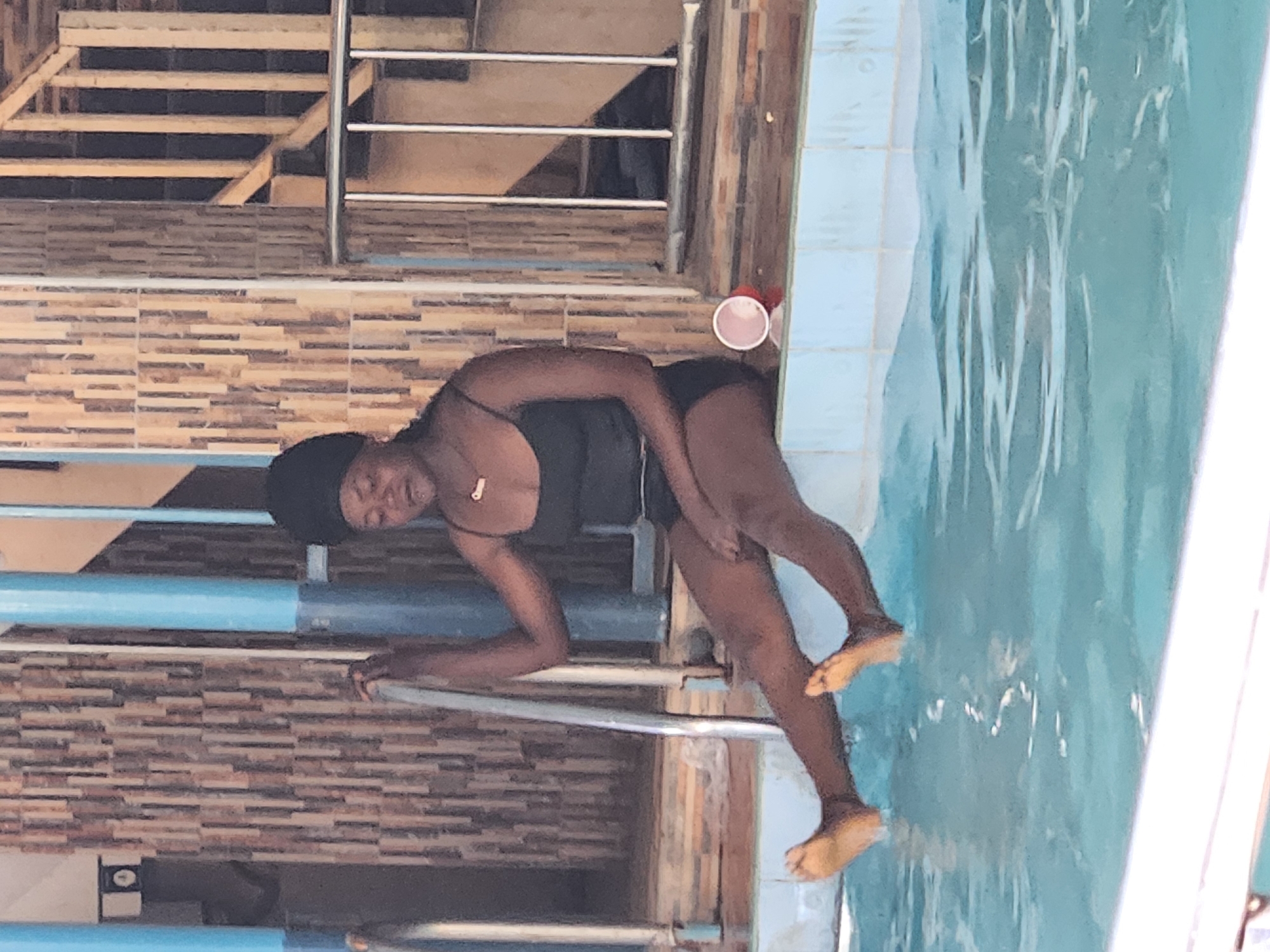 A woman wearing a black swimsuit and a head covering is at the edge of a swimming pool. She is partially in the water, holding onto a metal railing with her left hand. The pool has light blue tiles and clear water. Behind her is a tiled wall and a staircase with metal railings. A red and white cup sits on the edge of the pool near the wall. The scene is outdoors and well-lit.