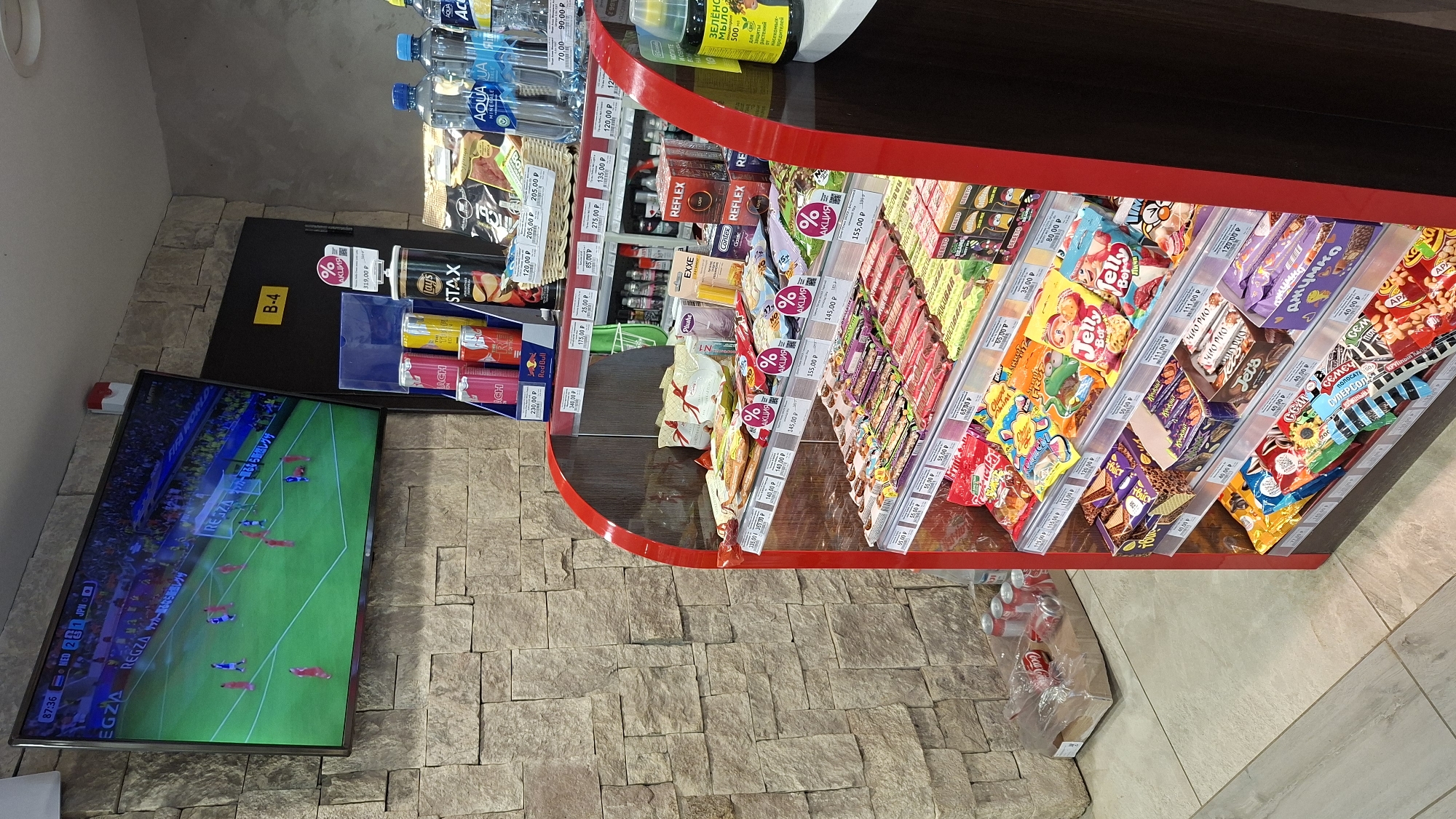 The image depicts the interior of a convenience store, likely in Nytva, Russia, judging by the Cyrillic text visible on the products. A large flat-screen television mounted on a stone-tiled wall displays a soccer match. To the right of the television, a door is marked with a yellow sign reading "B-4".
In the foreground, a refrigerated display case is filled with a variety of snacks, including candy bars, chips, and drinks. Several items are marked with "% АКЦИЯ" (meaning "SALE" or "PROMOTION" in Russian). Below the display case, on the floor, are several cans of Coca-Cola, some in a plastic bag, and a cardboard box. The store's flooring appears to be tiled. The overall lighting suggests an indoor environment, typical of a retail space. No people are visible in the frame.