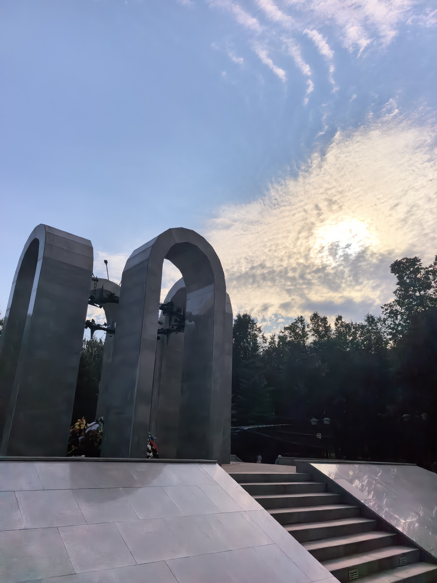 The image depicts a large, modern monument with several arched structures made of polished metal. The monument is situated outdoors, with a stone walkway and steps leading up to it. In the background, a dense forest of dark green trees lines the horizon. The sky above is a clear blue, with scattered white clouds illuminated by a bright sun peeking through them. There are no identifiable people clearly visible in the scene. The monument appears to be a place of remembrance, as a small arrangement of yellow and white flowers is visible at the base of one of the arches. The overall atmosphere is solemn and serene, with the natural elements complementing the monumental architecture. This monument is located in the city of Sterlitamak, Russia, and is part of the Eternal Flame memorial complex.