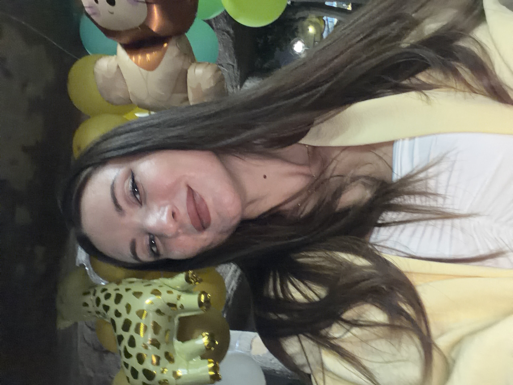 A selfie of a woman with long, dark hair is taken indoors, possibly at a party venue in 6th of October City, Egypt. She is wearing a yellow jacket over a white top and has a light amount of makeup. In the background, several balloons are visible, including a large giraffe-shaped balloon and a monkey-shaped balloon, suggesting a celebratory event, possibly a child's birthday party. The lighting is dim, with some ambient light sources present in the background, indicating the time of day is likely evening or night. The overall scene conveys a festive and informal atmosphere.
