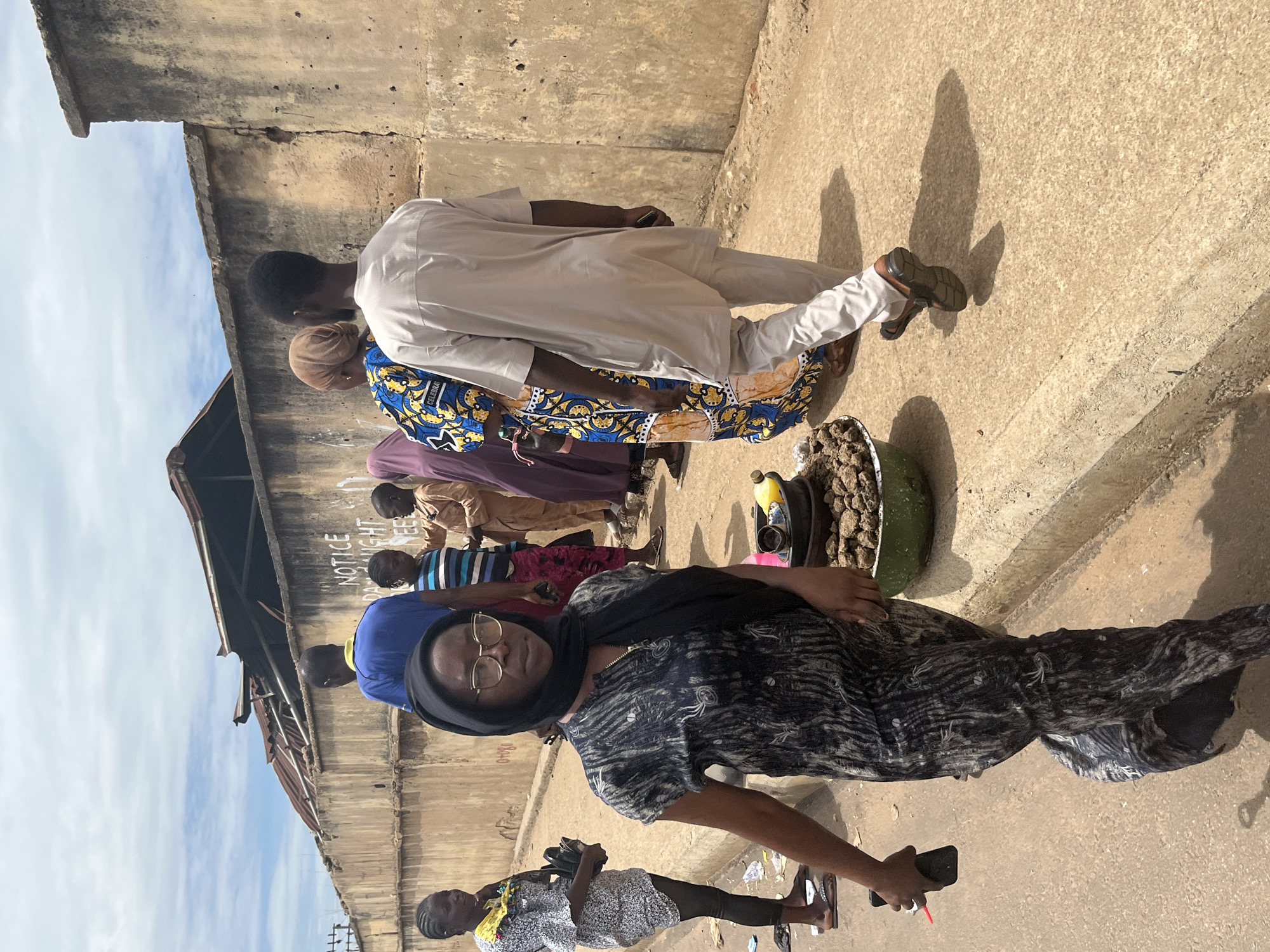 The image depicts an outdoor street scene in Jos, Nigeria, under a partly cloudy sky. In the immediate foreground, a woman with dark skin, wearing a black headscarf, gold-rimmed glasses, and a black and white patterned dress, walks towards the camera, holding a black smartphone with a red clip in her right hand. To her right, on the edge of the light-colored concrete sidewalk, rests a green basin containing a pile of dark, irregular-shaped objects, a small yellow bottle, and a dark cup, indicative of a street vendor's display.
Further back on the left, a woman in a light-patterned short-sleeved top and black leggings stands or walks, carrying a black bag. In the mid-ground, a man in a blue t-shirt and dark trousers walks away from the viewer, followed by several individuals positioned near a textured, light brown concrete wall. Visible black text on the wall partially reads "NOTICE DAYLIGHT VEE". Among these individuals are a person in a blue and white striped shirt and another in a light brown traditional garment. On the right, a woman in a brown head covering and a blue and yellow patterned dress walks beside a man in a light beige tunic and matching trousers, both moving away from the camera. The man wears dark sandals and holds a dark object in his left hand, casting a clear shadow on the ground.
Above the concrete wall, remnants of structures with dark, possibly charred, corrugated metal roofing are visible. The ground consists of a light-colored, dusty pavement, with some scattered litter near the curb.