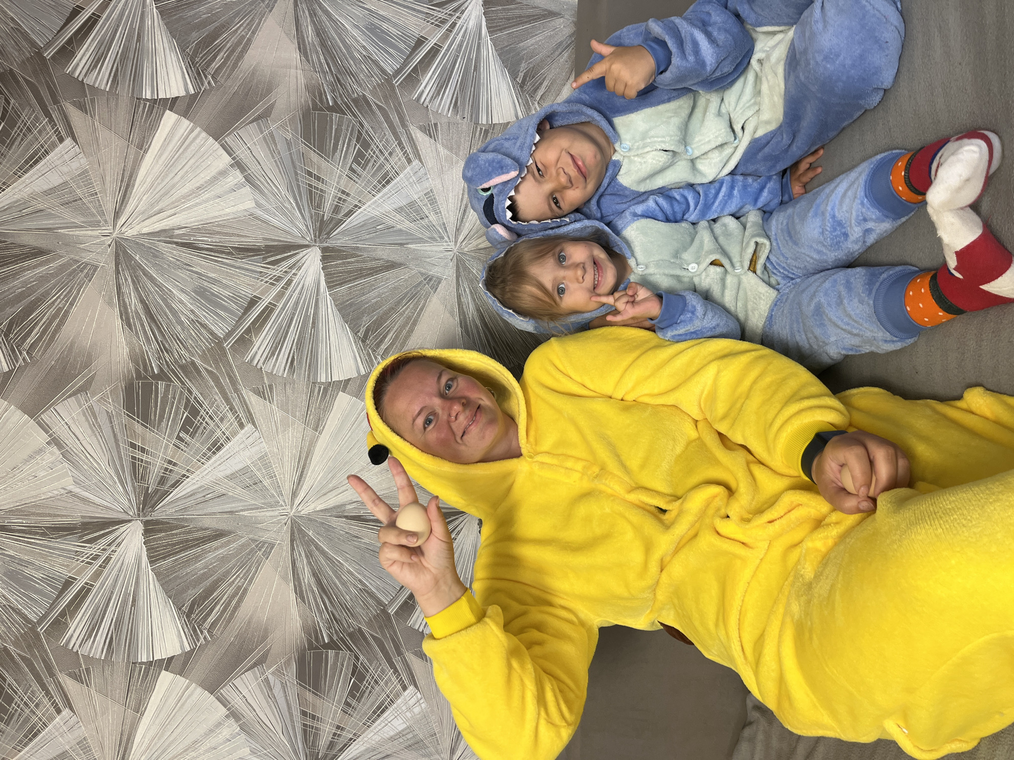 The image shows a woman and two children lying on a gray sofa in front of a wall with geometric patterned wallpaper. The woman is wearing a yellow Pikachu onesie and holding an egg in her left hand while making a peace sign. The two children are wearing blue Stitch onesies. The older child is pointing with his right hand. The location is Chaikovsky, Russia.