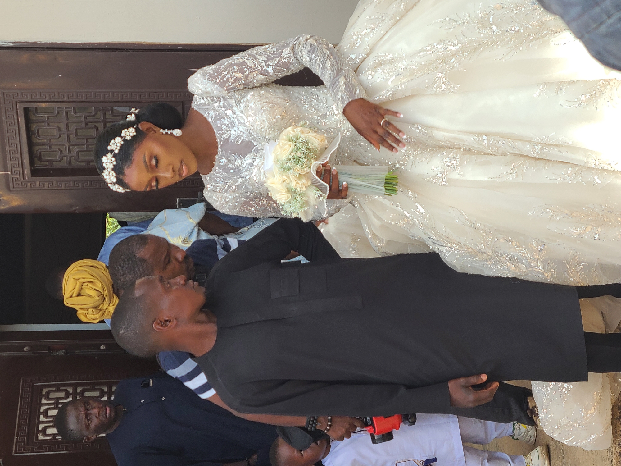 A bride in a white, long-sleeved wedding dress holding a bouquet of white roses stands surrounded by people. A man in a black suit is positioned closely beside the bride. The location appears to be outdoors, possibly near a building or structure. This scene is observed in what is reportedly the city of Barakin Lerawi, Nigeria.