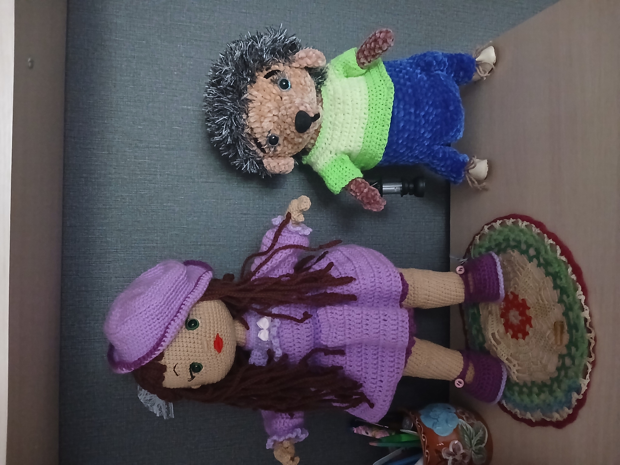 This is a photograph of two handmade stuffed toys on a shelf. On the left is a crocheted doll with brown yarn hair, green button eyes, and red lips, wearing a purple dress and a matching hat. On the right is a crocheted hedgehog toy with fuzzy grey and black yarn for its quills, wearing a green and light green striped shirt and blue pants. Both toys are standing on a wooden surface, and the background is a grey textured wall. To the left of the doll is a brown ceramic pot with colorful designs, filled with writing utensils. The overall scene is indoors and appears to be in a domestic setting. The location in the city of Климовское, Russia, is not discernible from the image itself.