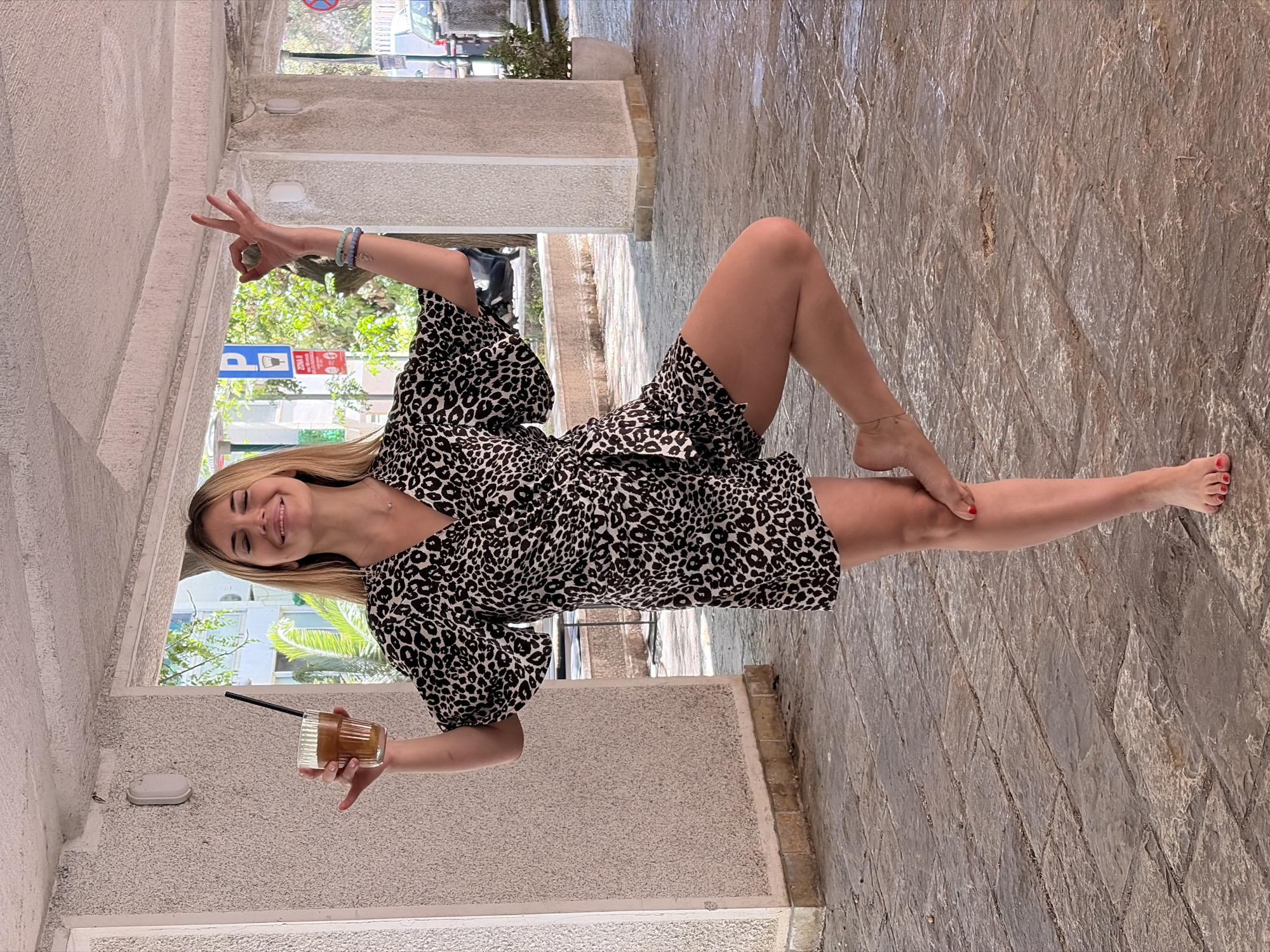 A fair-skinned woman is situated in an outdoor location, potentially within the city of Budva, Montenegro. She wears a black and white patterned dress with puffed sleeves. Her blonde hair is shoulder-length. She is holding a glass with a straw in her left hand. Her right hand is raised in a "peace" sign. She has an anklet on her right ankle. Her right leg extends out. The background suggests an urban environment, with architectural details reminiscent of Mediterranean structures. The ground appears to be paved with stone.