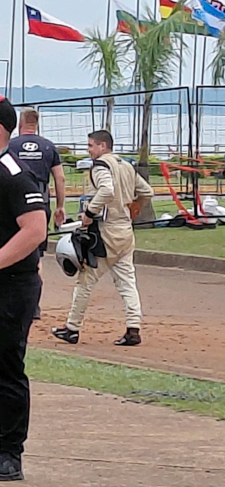 The image depicts an outdoor scene likely at a sporting event, possibly a race, in Encarnación, Paraguay. The primary subject is a man in a light-colored racing suit, walking towards the right of the frame. He is holding a white helmet with black straps in his left hand and has his right hand tucked into the back pocket of his suit. He is wearing black racing boots. To the left of the frame, another person is visible, wearing a black shirt and black pants, also facing towards the right. In the background, several flags are displayed, including a Chilean flag. Palm trees and fencing are also visible, suggesting a warm climate. The ground appears to be a mixture of dirt and grass.