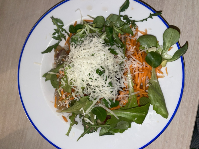 The image displays an overhead view of a prepared salad situated on a white circular plate featuring a blue band along its inner rim. The plate is positioned on a light-colored surface with a visible horizontal grain. The salad consists of a substantial central topping of finely grated white cheese. Beneath and around this cheese are julienned orange carrots and various green leafy vegetables, specifically identifiable arugula with its serrated edges and smaller, rounded leaves. A darker, somewhat translucent layer is discernable beneath the primary salad components. No individuals are present, and no actions are being performed. The environment suggests an indoor setting. The scene is located in Boreti, Montenegro.