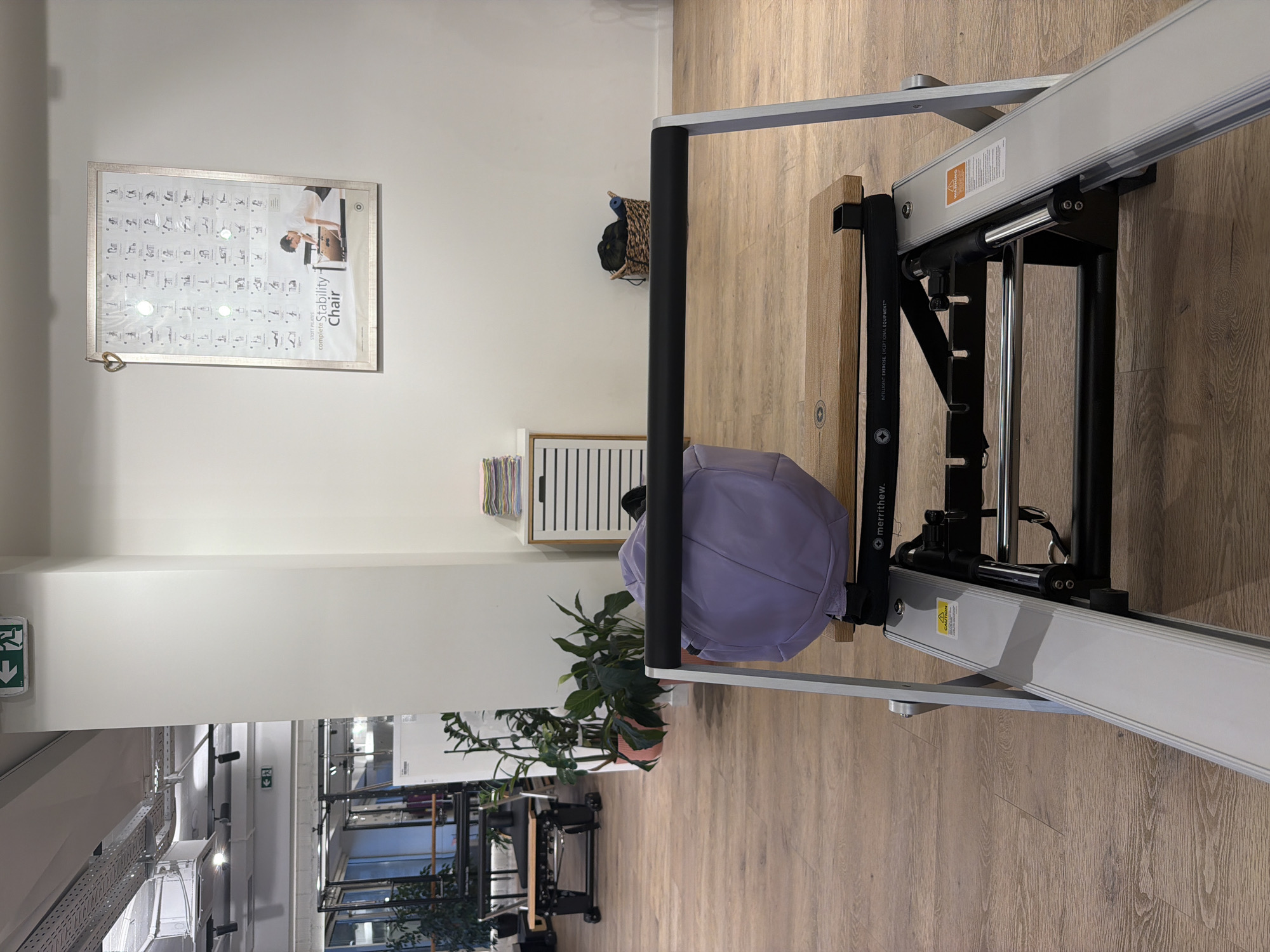 An indoor scene in what appears to be a Pilates studio. A reformer machine is in the foreground, with its carriage and wooden platform visible. A light purple exercise ball rests on the platform. Behind the reformer, the room features wooden floors, white walls, and large windows. Several Pilates apparatus are visible in the background, along with plants, a storage cabinet with stacked towels, and a framed poster on the wall detailing Pilates exercises on a stability chair. A woven basket containing rolled mats or towels sits on the floor. The lighting suggests an interior space with overhead fixtures. There are no people present in the image.