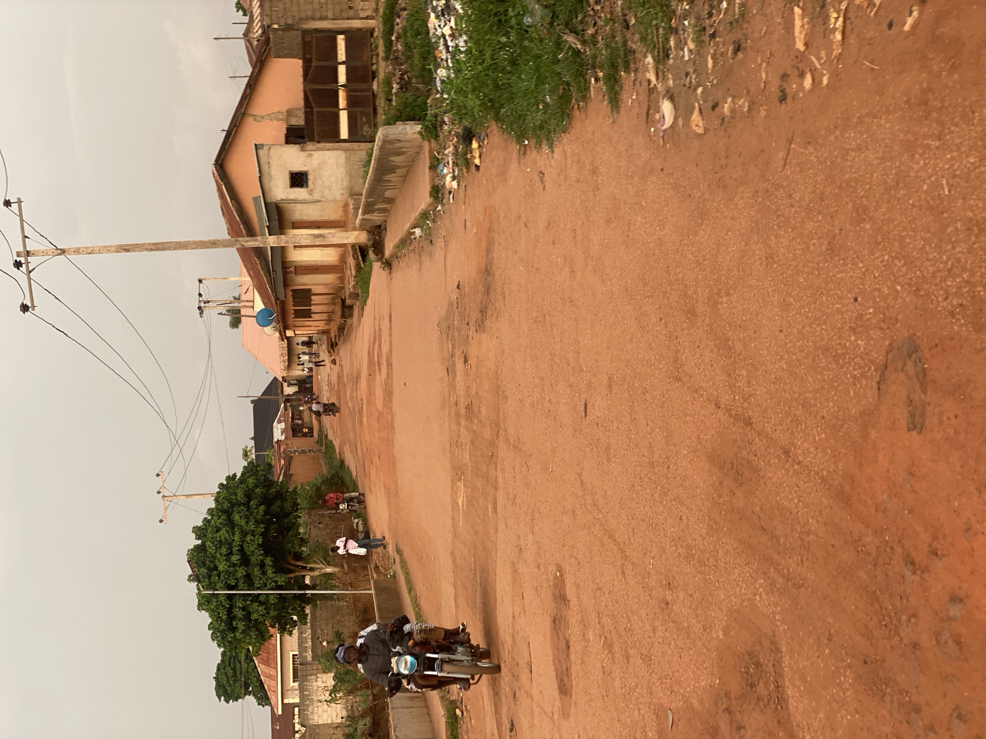 The image shows a dirt road in a residential area, likely Barden Yana, Nigeria. The road is reddish-brown in color and appears unpaved, with some loose stones and dust. On either side of the road are houses with corrugated iron roofs and concrete walls. There are also some trees and vegetation, and evidence of trash accumulation along the roadside.
In the foreground, two people are riding a motorcycle. The driver is wearing a dark jacket and a beanie, and the passenger is wearing a light-colored shirt. They are moving away from the camera, down the road. In the midground, there are a few other individuals walking and standing near the houses. Another motorcycle is parked near one of the houses. In the background, more houses and trees are visible, along with overhead power lines supported by wooden poles. The sky is overcast, suggesting a possibly cloudy or hazy day. The overall scene depicts a typical street view in a developing urban or semi-urban environment.
