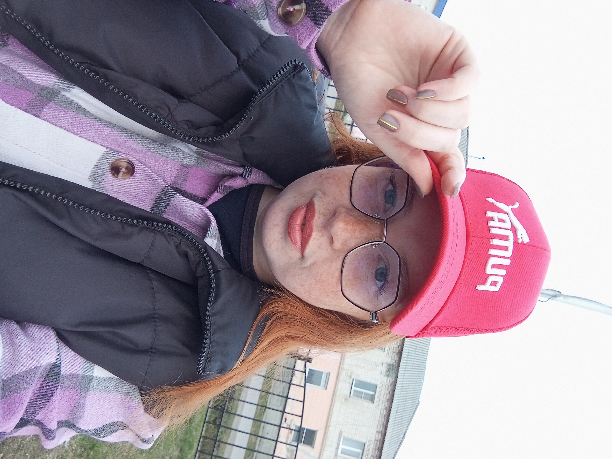 A young woman is captured in a close-up, slightly tilted portrait, seemingly a selfie, in Yurgamysh, Russia. She has reddish-auburn hair, fair skin with noticeable freckles, and light-colored eyes, which are visible through her thin-framed, hexagonal glasses. Her lips are painted a muted pink or coral shade.
She is dressed in a black zippered puffer jacket or vest worn over a plaid shirt featuring purple, white, and grey checks. A brown button is visible on her shirt. Underneath, a black top is peeking out. She is wearing a red baseball cap backwards, holding its brim with her right hand. Her fingernails are painted a muted taupe or grey color, each adorned with a thin, horizontal gold stripe.
The white text on the red cap, when viewed by the camera, reads "бМУЯ" in Cyrillic letters, appearing reversed and partially upside down due to the cap being worn backwards.
The background shows an outdoor setting with a light-colored brick building featuring several windows, likely residential or institutional. A metal fence or railing is visible between the subject and the building, with a patch of green grass at the bottom. The lighting is bright and diffused, indicating an overcast day during daylight hours. The overall impression is casual and relaxed.