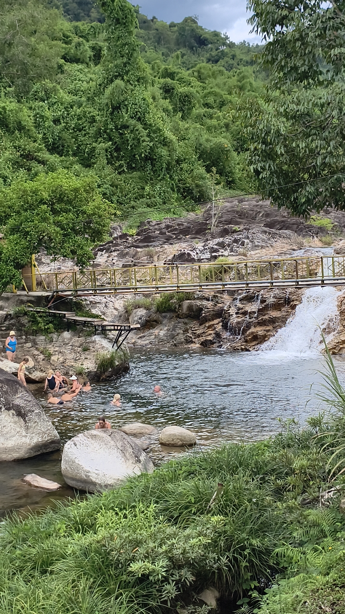 This is a vertical, medium shot photograph of a natural swimming area with a waterfall and a bridge, located in the city of Khanh Vinh, Vietnam. In the foreground, lush green grass and foliage grow along the bank of a calm body of water, with several large, smooth grey rocks scattered throughout the scene. A group of people are visible in the water, some swimming, others relaxing. To the left, two women are standing on a rock ledge, one wearing a blue swimsuit and the other a yellow bikini. A narrow, weathered wooden bridge with a yellow railing spans across the water and rocks in the mid-ground, leading to a small, cascading waterfall on the right side of the image. The background is dominated by a steep, densely forested hillside covered in a variety of green trees and vegetation. The sky above is overcast with a soft, diffused light.