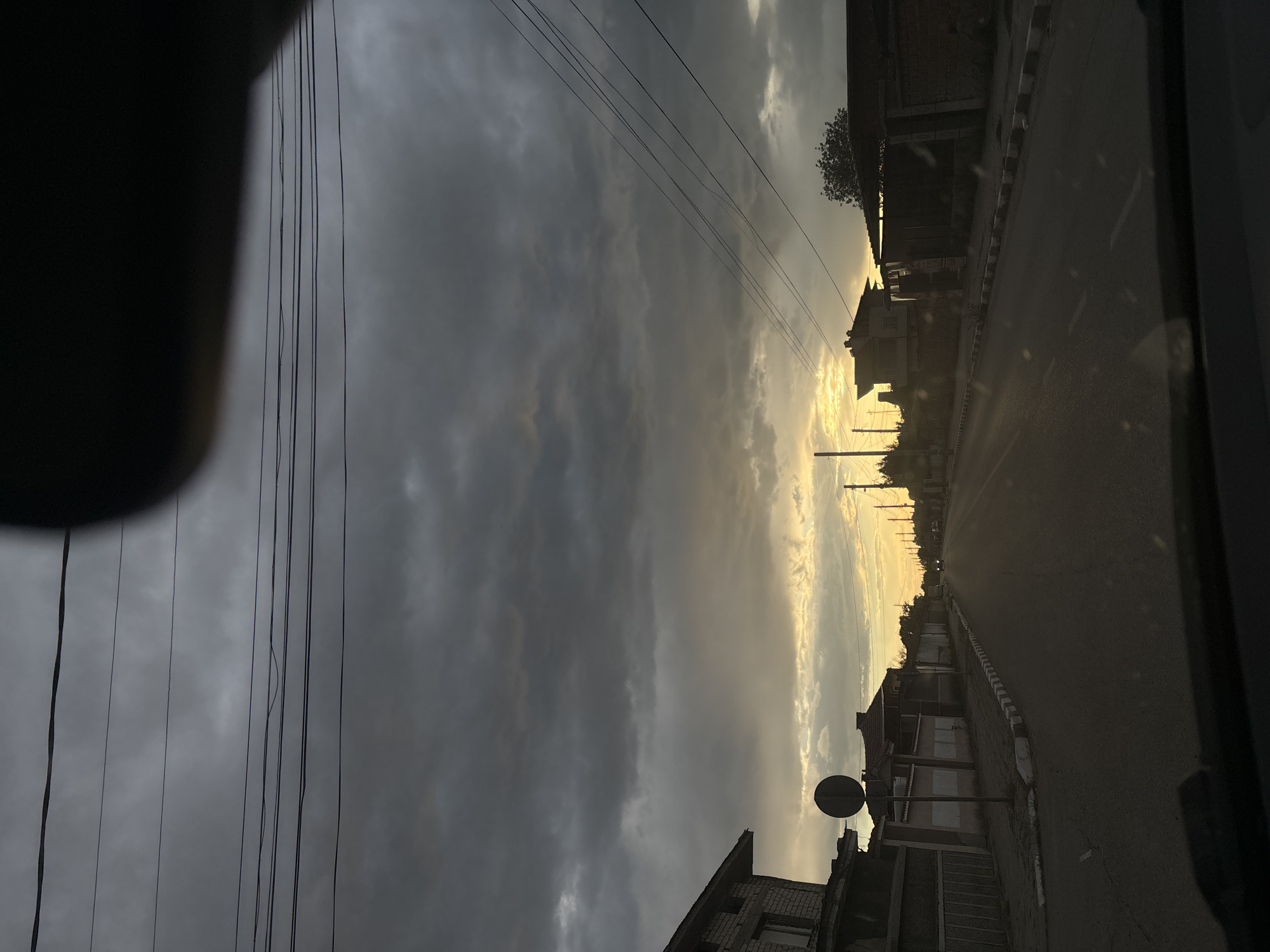 The image shows a street view in Manole, Bulgaria, at dusk. The sky is filled with dark, overcast clouds, with a strip of yellow light visible towards the horizon, indicating a setting sun. Utility lines are strung across the sky. The road stretches into the distance, lined with buildings on both sides. A road sign is visible on the left side of the road. The view is partially obstructed by a dark object on the left, possibly a part of the vehicle from which the image was taken. The overall mood is somber, reflecting the heavy clouds and low light.