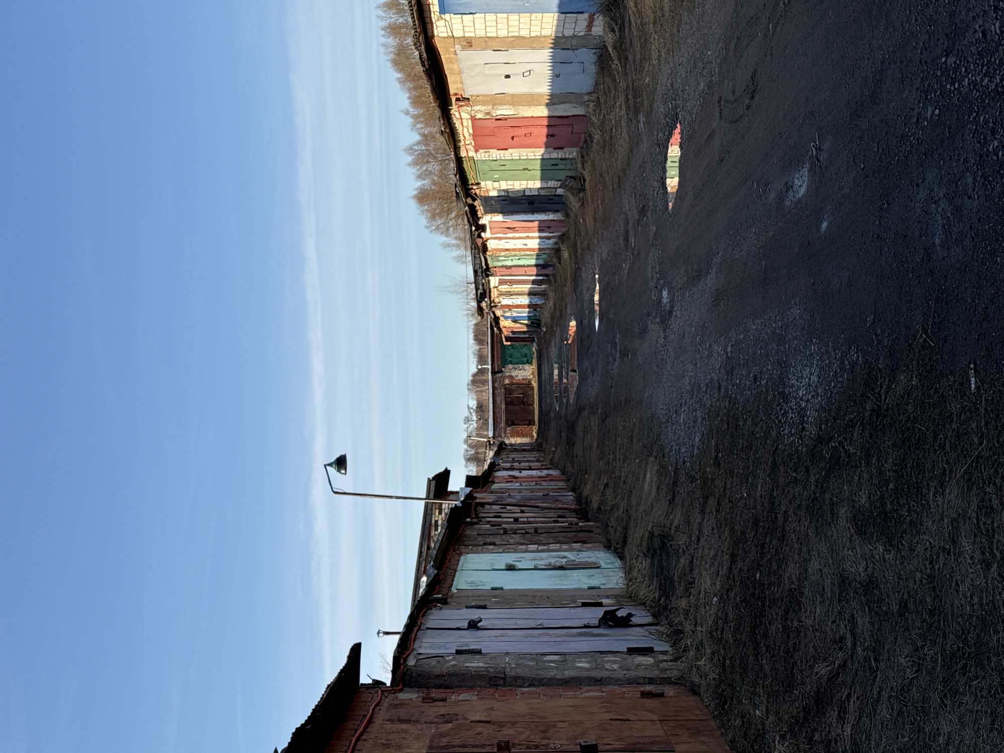 The image depicts a narrow alleyway between two rows of garages in Izhevsk, Russia. The garages are constructed from brick and concrete, with painted metal doors in varying colors. The alleyway's surface is asphalt, showing signs of wear and disrepair with puddles and patches of bare ground. Overhead, a lone street lamp is present. The sky above is blue with some clouds. There is no evidence of people or activity.