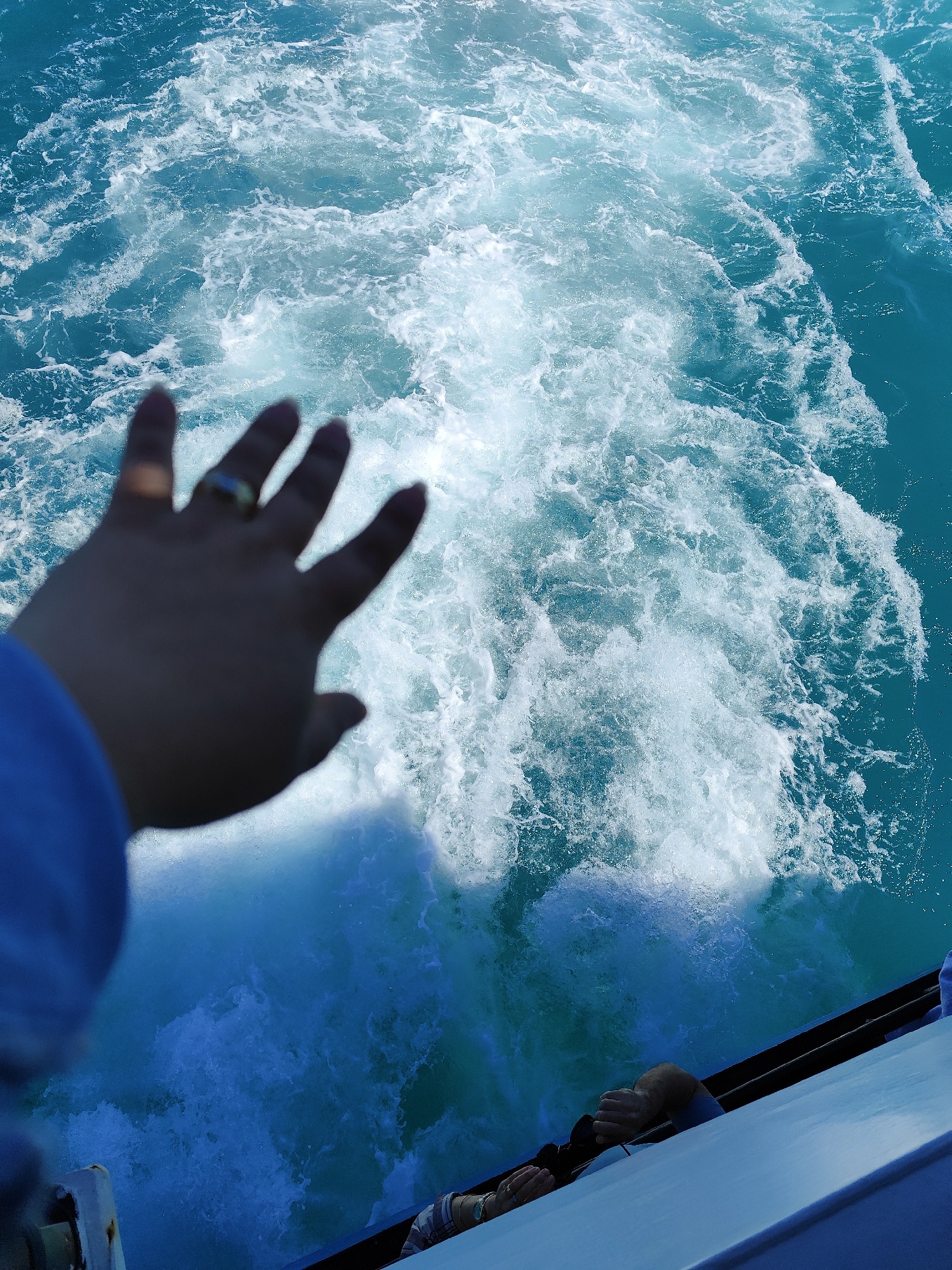 A view of turbulent, turquoise water churned by a boat is presented from a high vantage point, likely on the boat itself. A person's left hand is prominently displayed in the foreground, fingers outstretched toward the water, a ring visible on the ring finger. The back of another person's head and hands are barely visible at the very bottom of the frame, suggesting they are sitting or leaning over the side. The image's location is Sarıyer, Turkey, inferred from the water color and boat context, typical of the Bosphorus Strait. The environment consists of the open sea and the vessel's edge. The boat is moving, creating the wake of foamy water in its path.