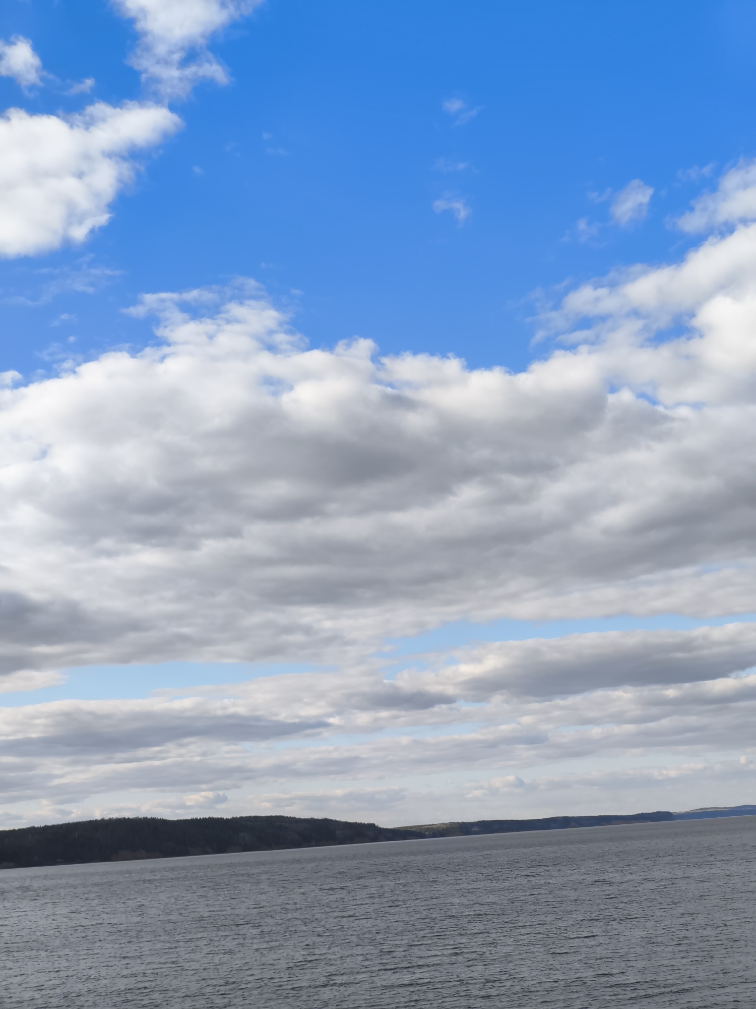 The image depicts a body of water, likely a river or a large lake, with a treeline of hills or low mountains in the distance. The water is rippled, suggesting a slight breeze. The sky dominates the upper portion of the image, featuring a mix of blue patches and layered clouds of varying density. Some clouds are bright white, while others are darker and grey, indicating potential overcast conditions.
Without additional landmarks or identifying information, definitively placing this image in the city of Chaikovsky, Russia, is impossible.