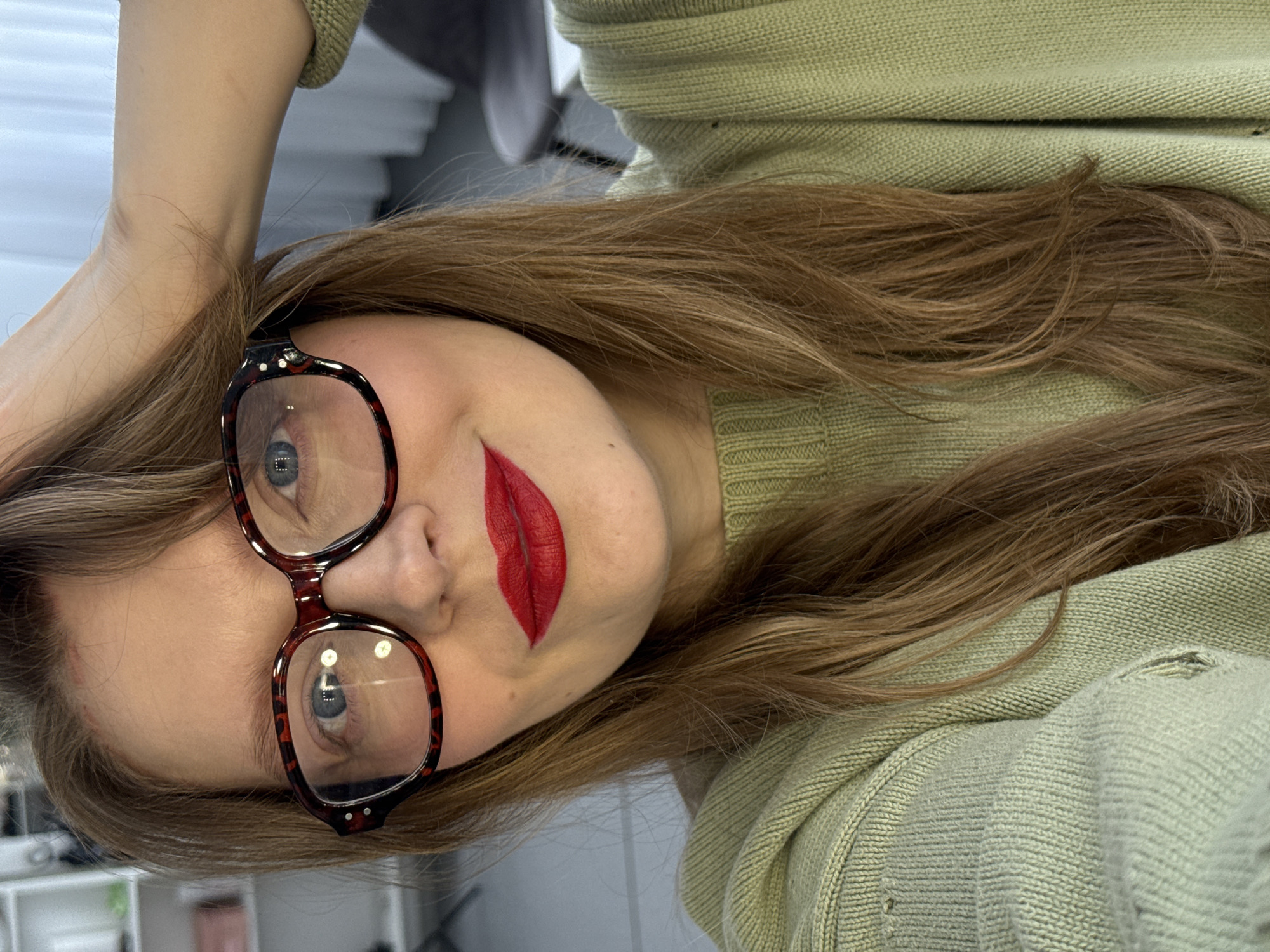A woman with long brown hair and blue eyes is posing for a selfie. She is wearing glasses with a brown and red patterned frame. Her lips are painted with bright red lipstick. She is wearing a light green sweater. The background appears to be an indoor setting, possibly a home or office, with neutral colors and minimal clutter. There is no clear indication of Izhevsk, Russia in the image.