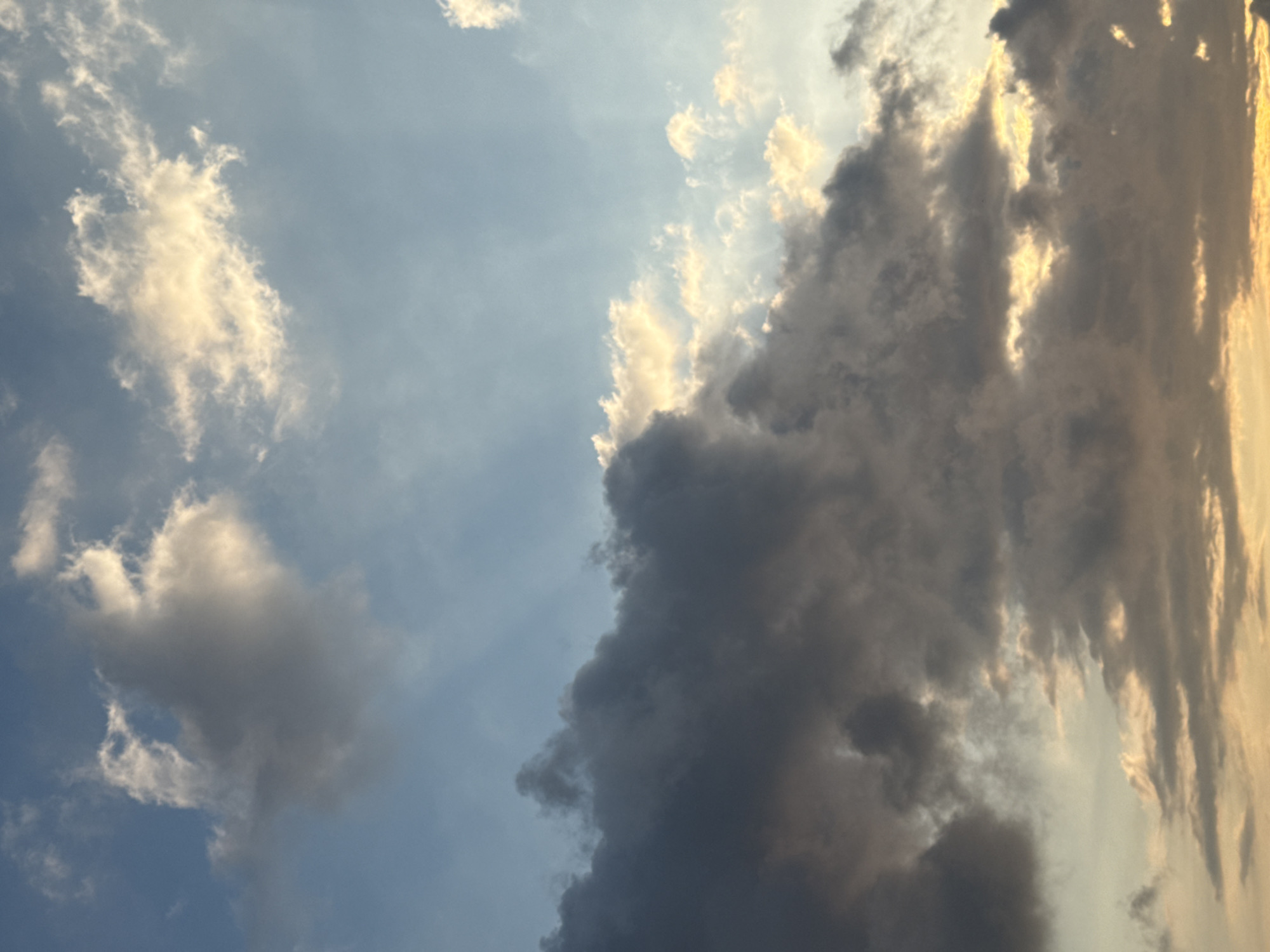 The image depicts a sky filled with a mix of dark gray and bright white clouds. Sunlight is breaking through some of the clouds, creating visible rays. The sky is a gradient of light blue to a warmer hue near the horizon, suggesting either sunrise or sunset. There are no visible people, structures, or any other identifiable objects or subjects in the image; it is exclusively focused on the atmospheric conditions of the sky. The location is unknown.