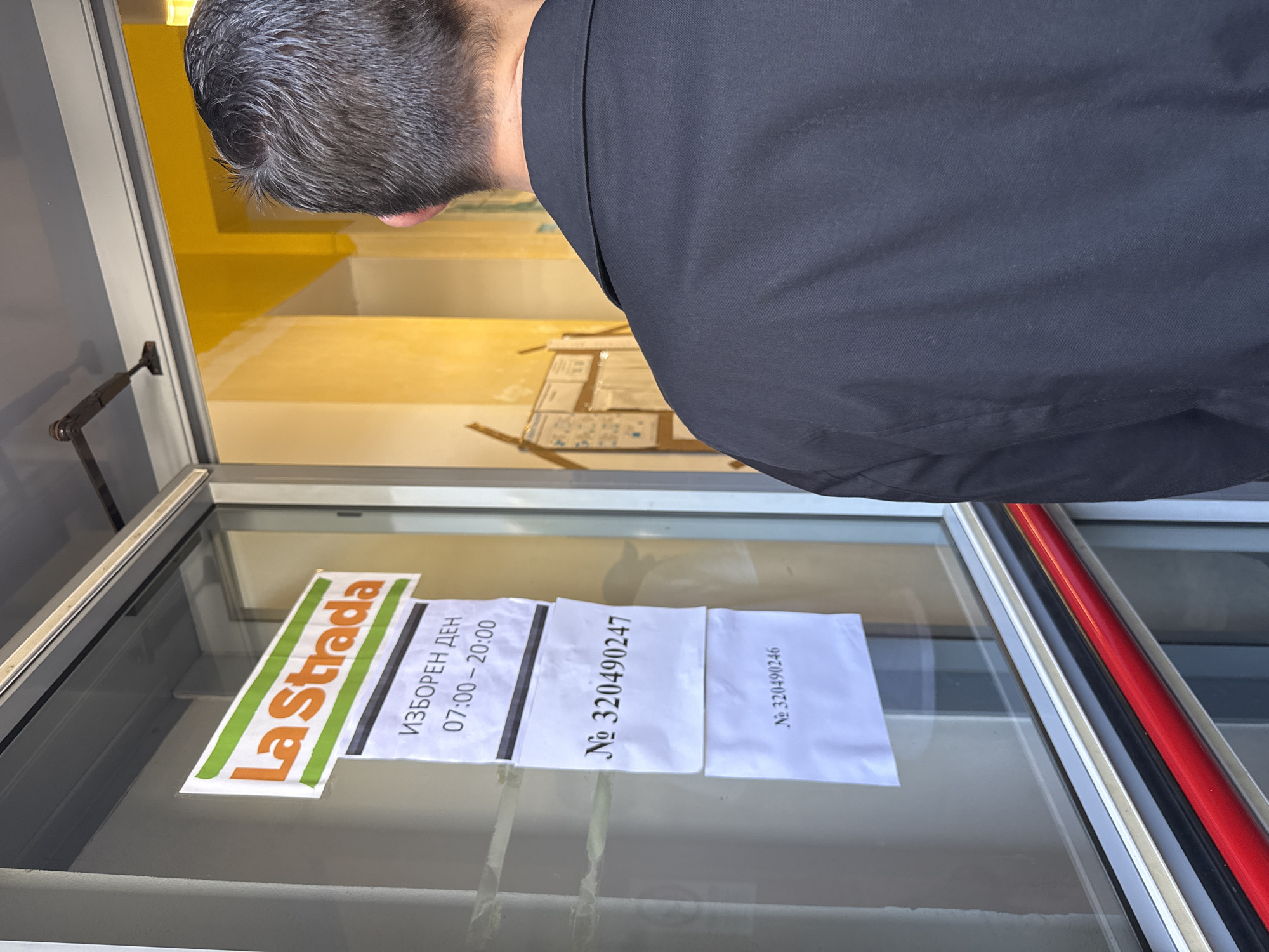 A man with grey hair is seen looking into the display window of La Strada, a business in Milan, Italy. Affixed to the inside of the window are papers with the following: a sign with the business name "La Strada"; another sign stating "Election Day" with hours 07:00-20:00; and two more signs that say "No 320490247" and "No 320490246". The interior of the business is visible through the window.