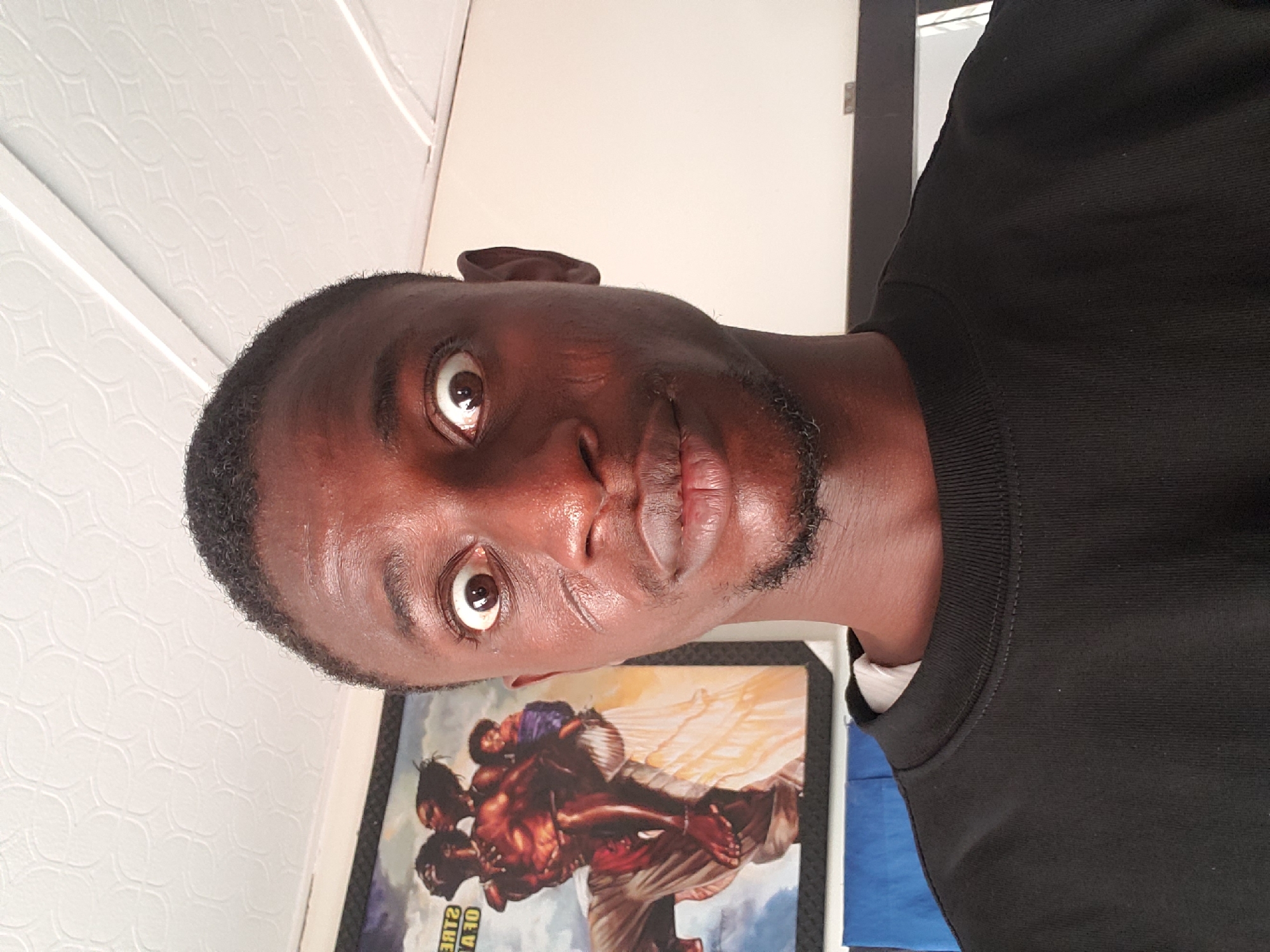 The image presents a close-up portrait of a dark-skinned individual, likely male, whose head is tilted, as the original photograph is rotated 90 degrees counter-clockwise. He has short, dark, closely-cropped hair and sparse facial hair along his jawline. His eyes are wide open, with the whites prominently visible, as he looks directly at the camera, conveying an alert, intense, or possibly surprised expression. His lips are slightly parted. There are reflections on his forehead and lips, suggesting natural light or a light source shining on his skin.
He is dressed in a plain black long-sleeved shirt or sweatshirt, with a glimpse of a white undershirt visible at the collar. The black garment features a subtle ribbed texture around the neckline area.
The background consists of light-colored walls. To the left of the subject, there is a white or off-white wall with a repeating embossed geometric pattern. To the right, the wall is smooth and unpatterned. Partially visible below the person's head is a framed piece of artwork or a poster. This artwork features several muscular, dark-skinned figures in what appears to be a dynamic or dramatic scene, with at least one figure carrying another on their back, set against a backdrop of bright light or clouds. Inverted text on the artwork or its frame includes fragments such as "ZLBE" and "VA O".
The scene appears to be indoors in Adankolo, Nigeria, under clear ambient lighting.