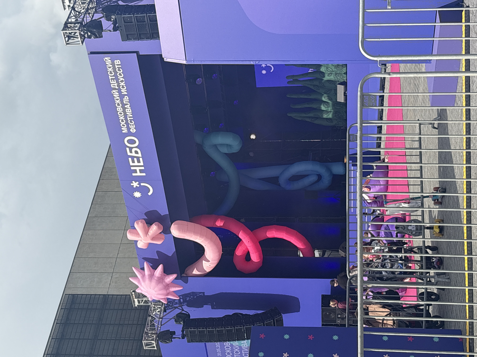 The image shows an outdoor stage in Moscow, Russia, decorated for the Moscow International Children’s Arts Festival "Nebo." The stage is predominantly purple with large, colorful inflatable decorations in the shapes of stars, worms, and foliage. The stage has signage in Russian indicating the festival name. In front of the stage, a metal barrier separates the stage from the audience, some of whom are visible behind the barrier. The weather appears overcast. There are buildings in the background behind the stage.