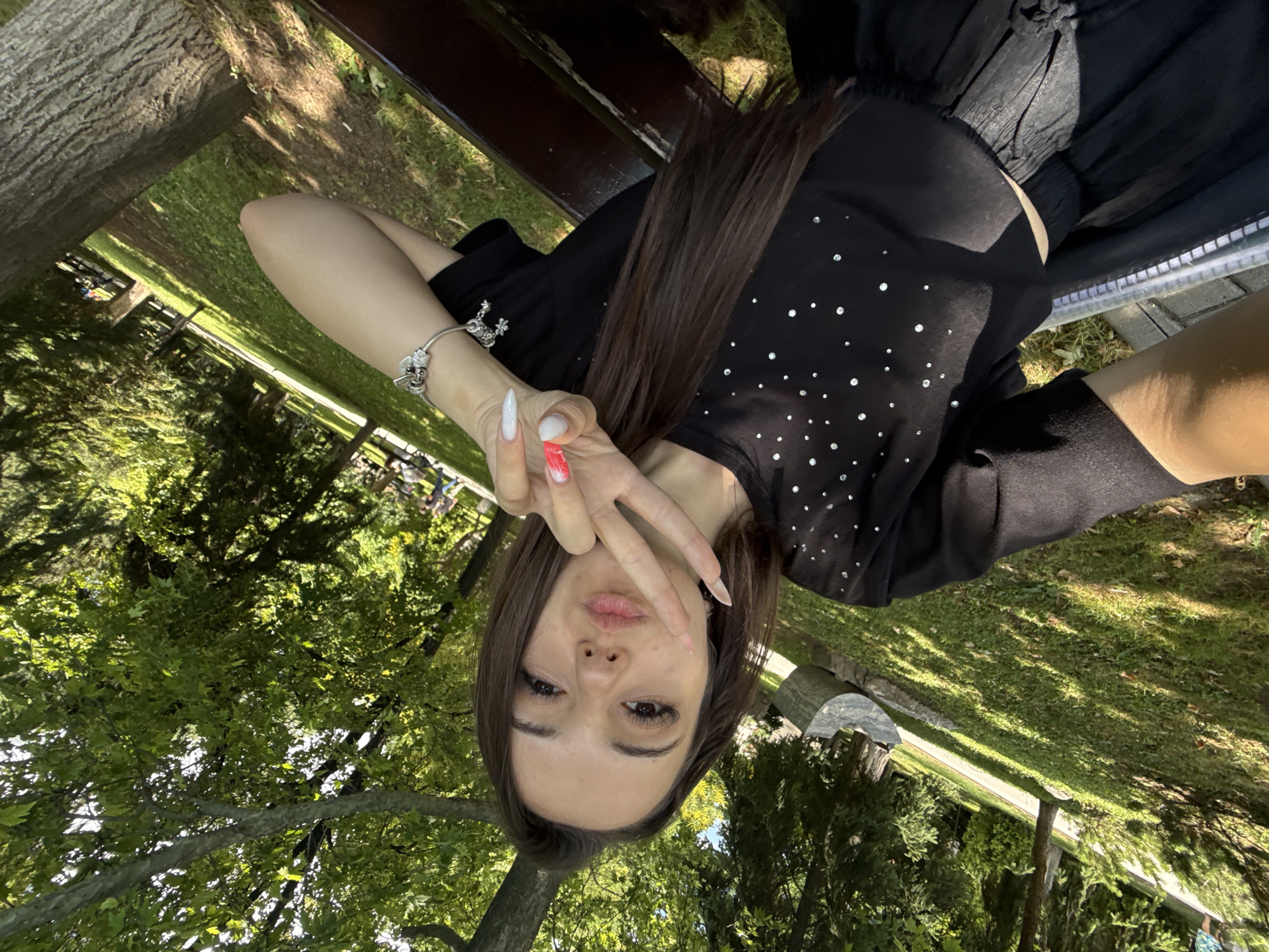 A young woman with long, dark brown hair and fair skin is taking a selfie outdoors, in a park. She is wearing a black t-shirt adorned with rhinestones and a black skirt. Her left arm is raised, and she is making a peace sign with her hand, displaying white and red-tipped fingernails. A silver charm bracelet is visible on her wrist. The background shows lush green grass, trees, and a wooden bench. The lighting suggests it is daytime, with sunlight dappling through the leaves. The orientation of the image is portrait. The scene appears to be a tranquil park setting, likely in the city of Plovdiv, Bulgaria, given the context.