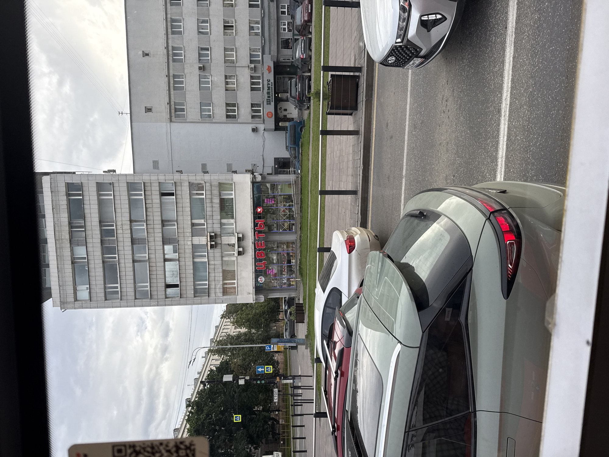 This image shows a street scene in Perm, Russia, with several cars parked along the side of the road. The cars are modern, with one silver car in the foreground and a white car behind it. In the background, there are several multi-story buildings, typical of urban architecture. One building has a sign that reads "ЦВЕТЫ" (Flowers) in red lettering, indicating a flower shop. The sky is overcast, suggesting cloudy weather. The scene appears to be during the daytime. There are also street signs and traffic lights visible, indicating this is a public street with traffic regulations. A QR code is visible in the bottom left corner of the image.