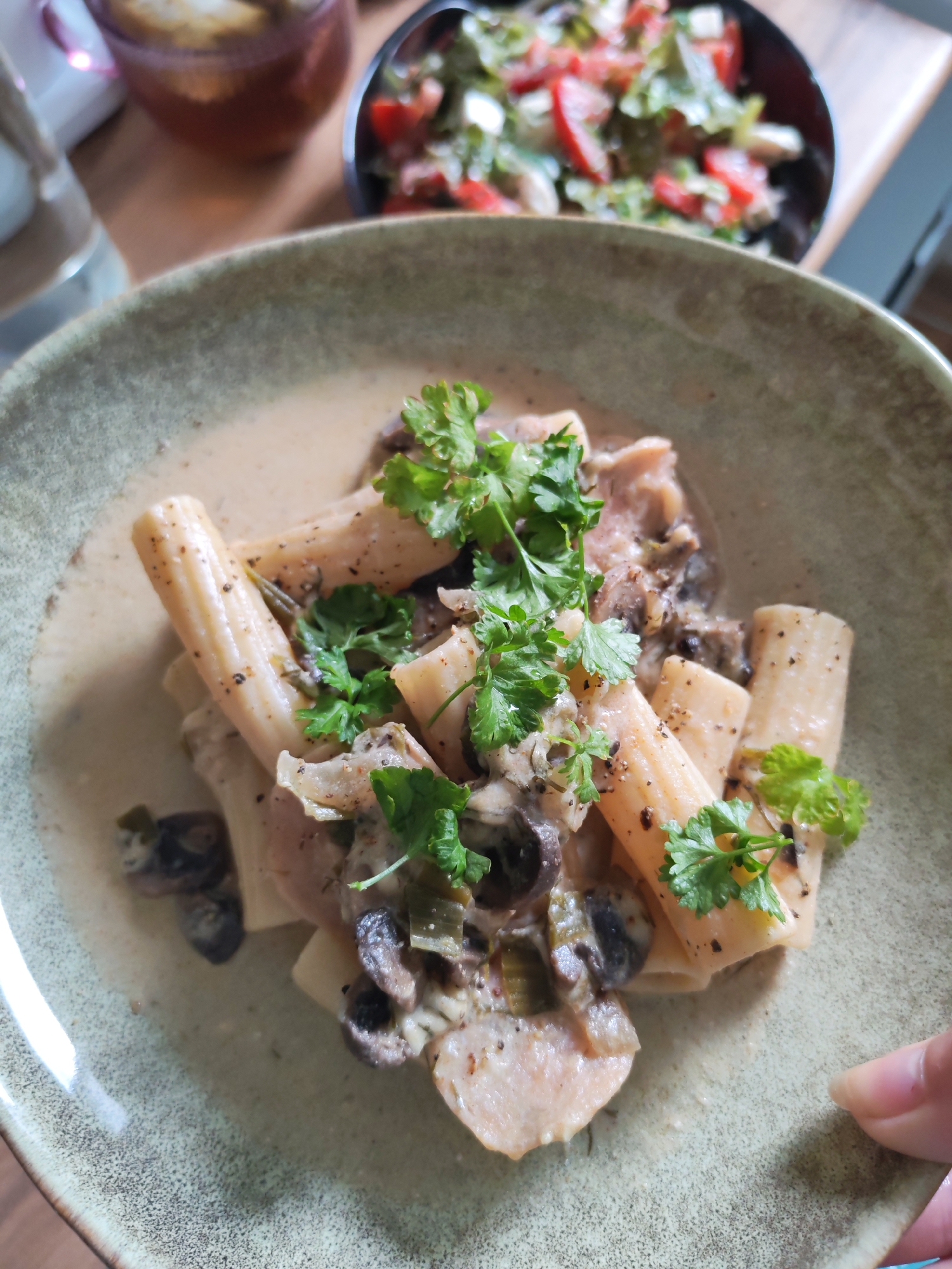 A close-up, overhead shot displays a bowl of pasta with a creamy sauce, garnished with fresh parsley and black pepper. The pasta appears to be rigatoni, and the sauce contains visible pieces of mushrooms and leeks. In the background, a blurry image shows a salad in a dark bowl and a glass of iced tea. The scene is set on a wooden table. There is no specific information that would indicate the location is Izhevsk, Russia.