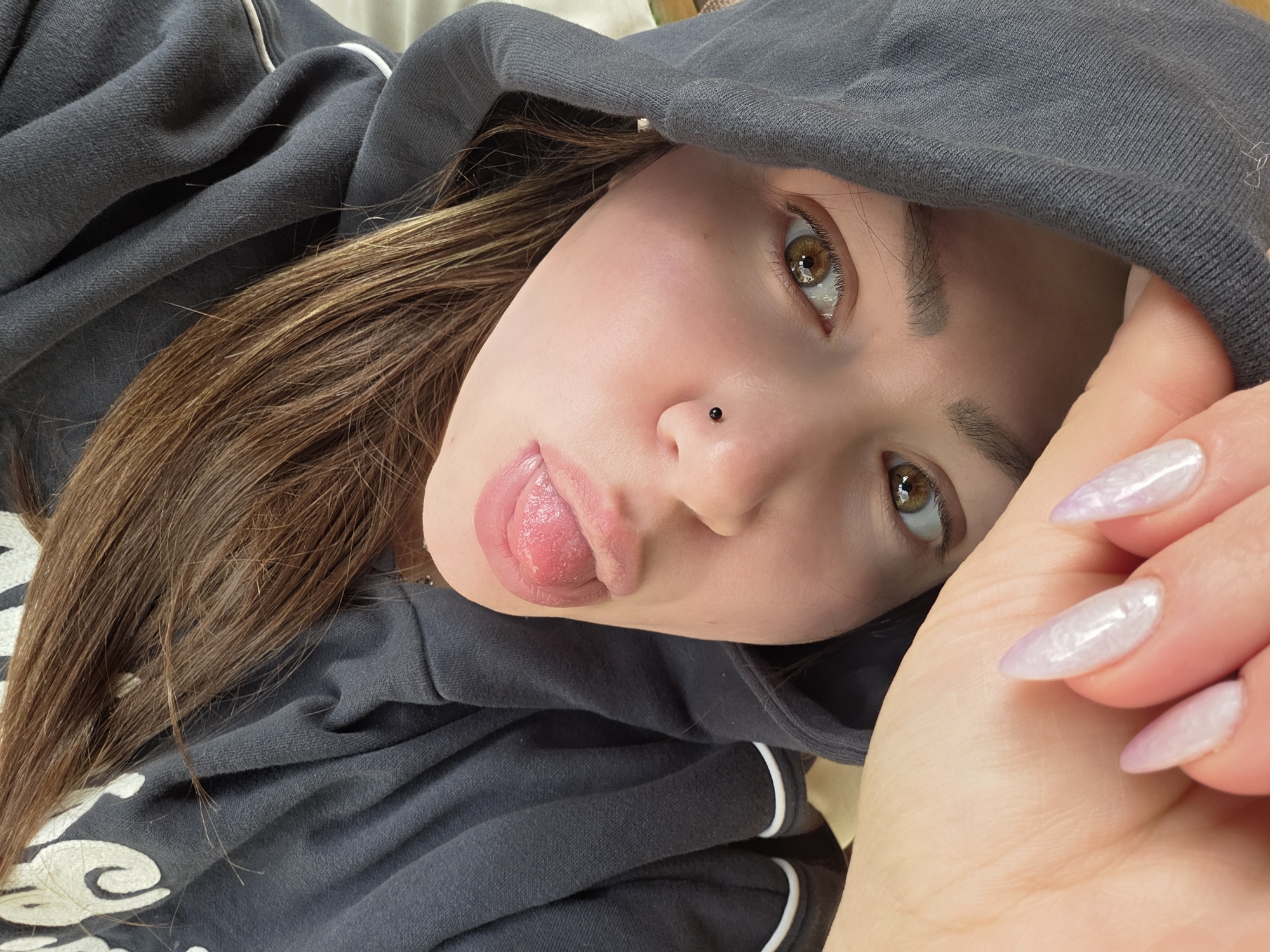 A close-up selfie shows a person, likely female, lying down or reclining. She looks directly at the camera with a playful expression, her tongue slightly extended. Her brown eyes are striking, and she has a small black stud piercing on her nose. Her long, light brown hair frames her face and spreads around her.
She is wearing a dark grey hooded sweatshirt with visible white piping detail along the edge of the hood and possibly seams. A small portion of a white design is visible on the front of the sweatshirt. Her left hand is prominent in the foreground on the right side, with long, almond-shaped fingernails painted in a shimmery, light lavender or iridescent polish. The background is soft and out of focus, suggesting an indoor setting. The image conveys a casual and relaxed mood. No text is visible within the frame, and there are no specific visual cues in the image to indicate the mentioned location of London, United Kingdom.