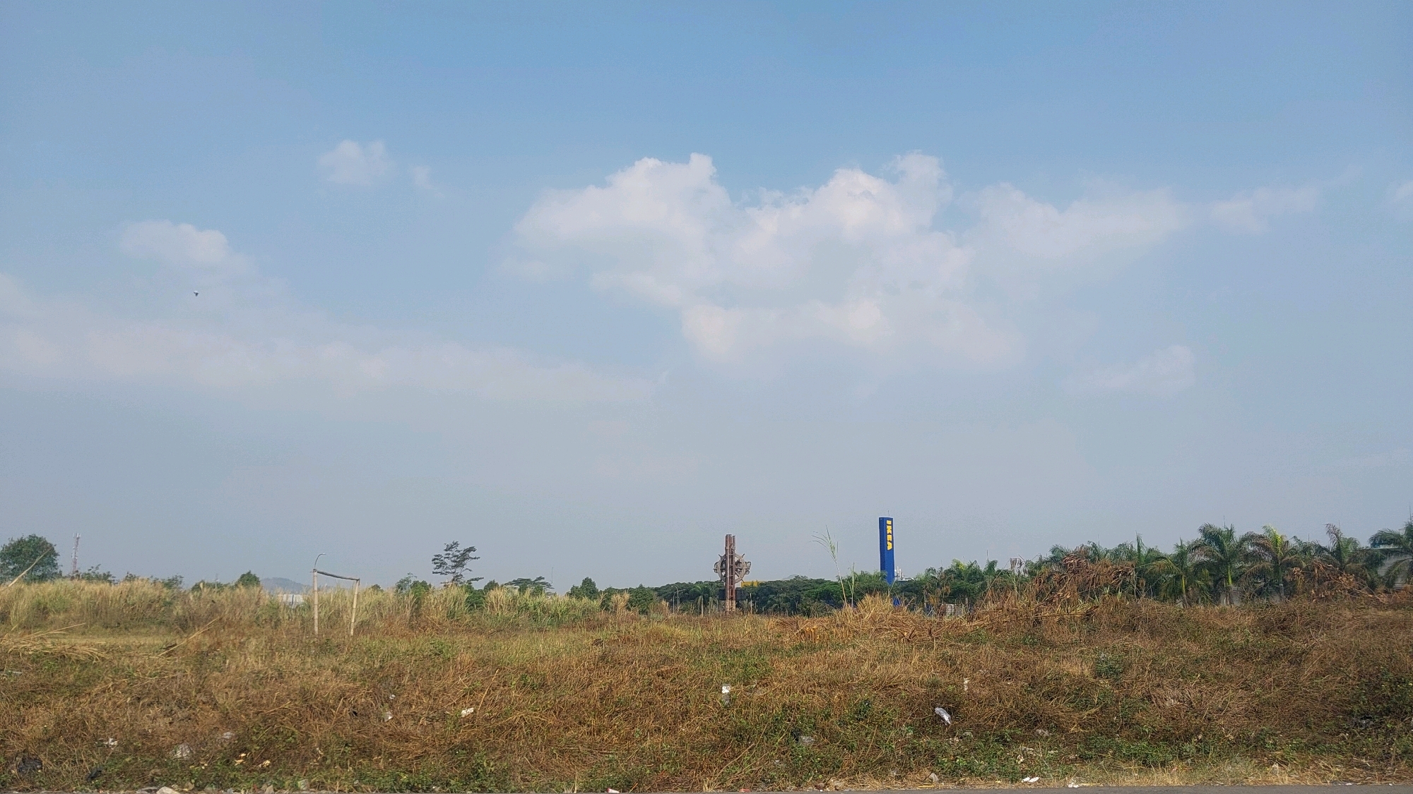 This is a landscape photograph taken in Jayamekar, Indonesia, under a clear blue sky with scattered clouds. The foreground is dominated by dry, brown grass and some scattered debris. In the midground, there is a field of tall, dry grass with a rudimentary wooden structure, possibly a goalpost, to the right of center. Beyond the field, a line of trees and palm trees forms the background. To the right of the frame, a tall blue sign with yellow lettering, which appears to be an IKEA logo, is visible. In the distance, a cell tower can be seen to the left of the frame. A small bird is flying in the upper left quadrant of the sky. The scene depicts an undeveloped or semi-developed area on the outskirts of the city.
