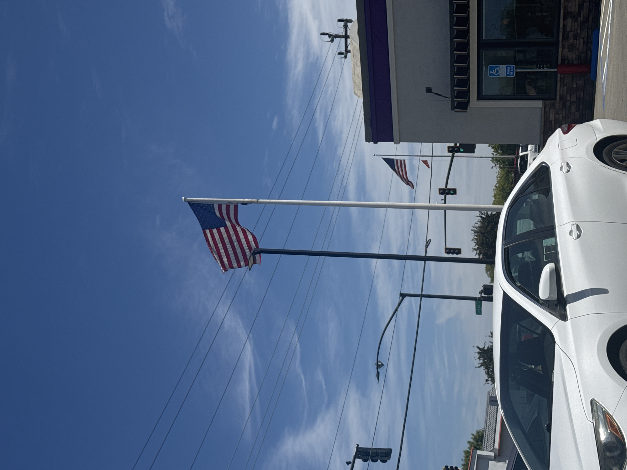 In Citrus Heights, California, a bright blue sky with thin white clouds forms the backdrop for a street scene. Two American flags are prominently displayed on flagpoles, one taller than the other. The taller flagpole, located on the left, is black and holds a large flag. The shorter flagpole, on the right, is white and holds a smaller flag. In the foreground, the front passenger side of a white sedan is visible, parked facing left. Behind the car, a building with a purple awning and a grey facade stands. A blue handicapped parking sign is posted near the building's entrance. Traffic lights and streetlights are also present, indicating an urban environment.