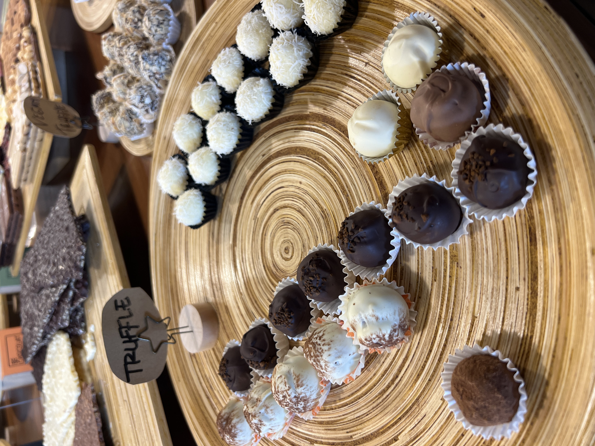 The image shows an assortment of chocolate truffles displayed on a wooden, spiraled-patterned platter. The truffles have different coatings including coconut flakes, plain white chocolate, milk chocolate, and dark chocolate with sprinkles. A small sign labeled "TRUFFLE" is visible near a tray of chocolate items. The location is in the city of Unknown, Unknown.