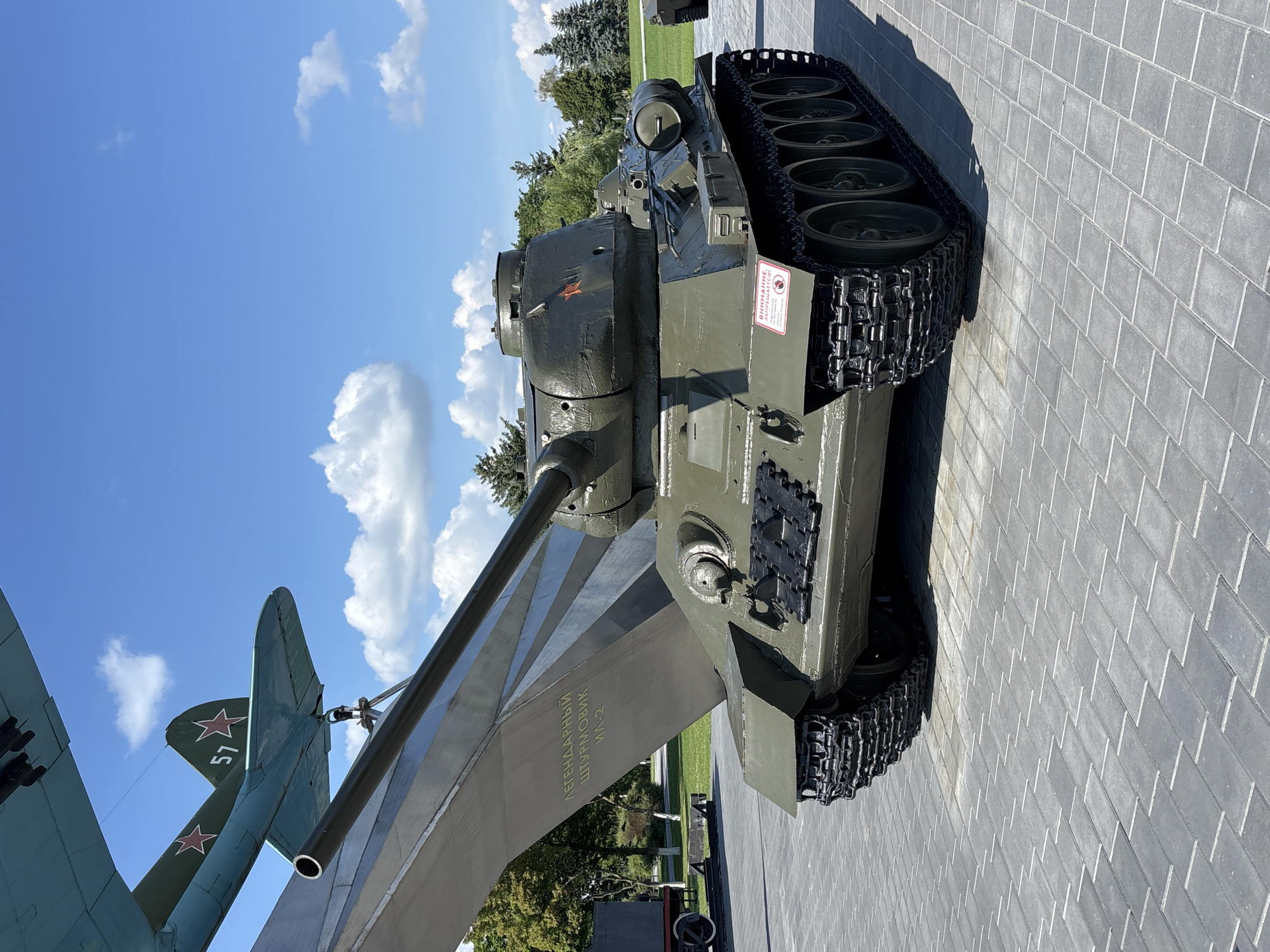 In the city of Ozyaritska-Slabadski rural council, Belarus, a military museum displays a Soviet-era tank, likely a T-34, painted in olive drab. The tank is positioned on a paved area with a distinctive herringbone pattern. Behind the tank, a large, metallic monument with engraved text rises, and a portion of a light blue airplane, possibly a Shturmovik Il-2, is visible at the top left, featuring a red star insignia and the number 57. The background consists of a clear blue sky with scattered white clouds, green trees, and a glimpse of another military vehicle to the right. A red warning sign is affixed to the side of the tank.