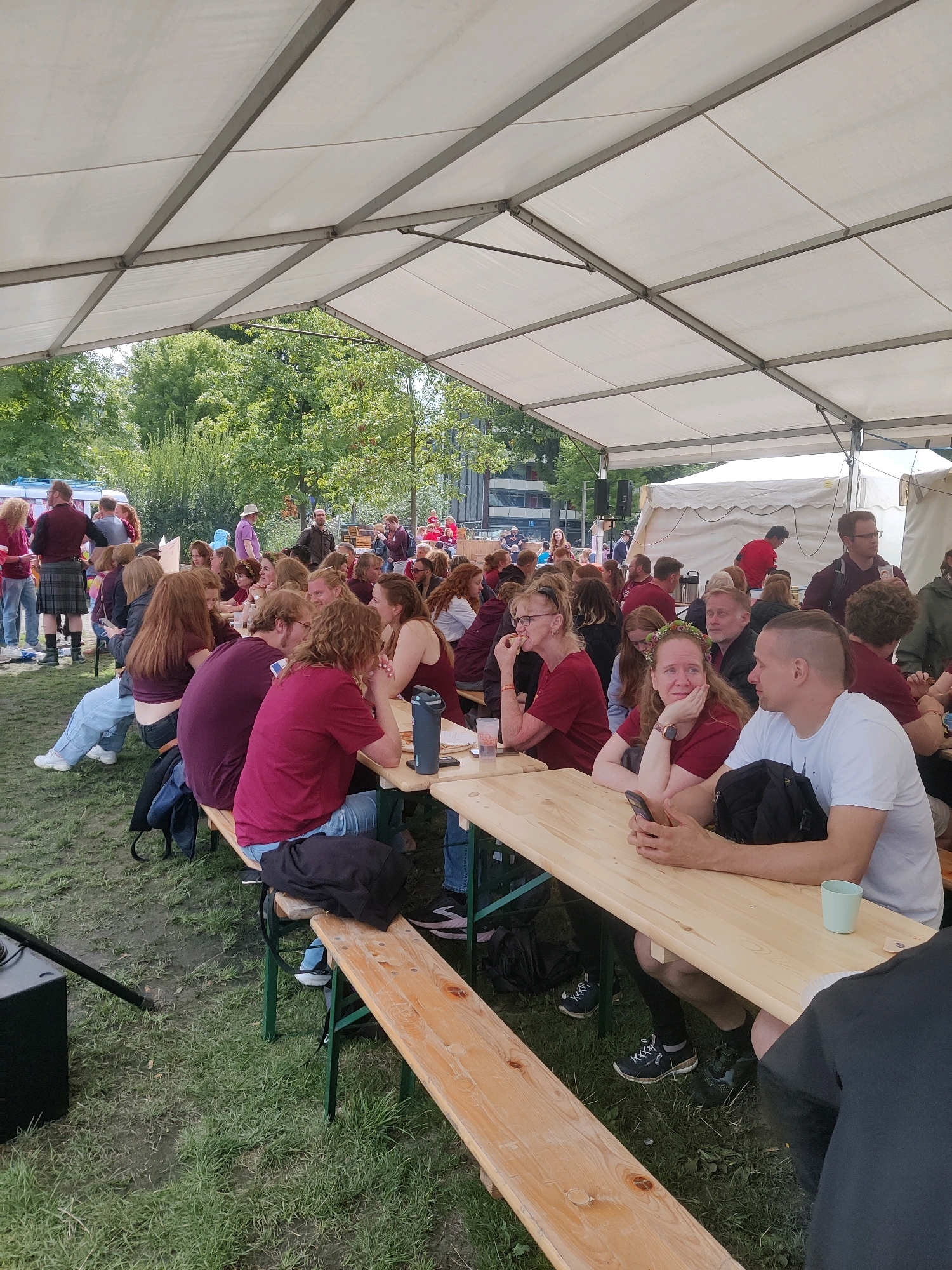 An outdoor event, likely a festival or market, is depicted under a large white tent structure. The event appears to be taking place in the city of Tilburg, Netherlands, based on the context of the original image source. A large gathering of people, predominantly young adults, are seated at long wooden picnic tables. Many are wearing casual clothing, with a noticeable prevalence of red and maroon t-shirts. Several individuals are engaged in conversation, while others are looking at their phones.
In the foreground, a man with a distinctive shaved haircut and a white t-shirt is seated next to a woman wearing a red t-shirt and a flower crown. They are interacting, and the man is holding a mobile phone. To their left, a black speaker is positioned on the ground next to a wooden bench. The ground is covered in grass and some areas appear to be muddy.
In the background, more people are seated at tables, and further back, a white tent building is visible. Trees and some buildings can be seen beyond the tent. The overall atmosphere suggests a social and informal gathering.
