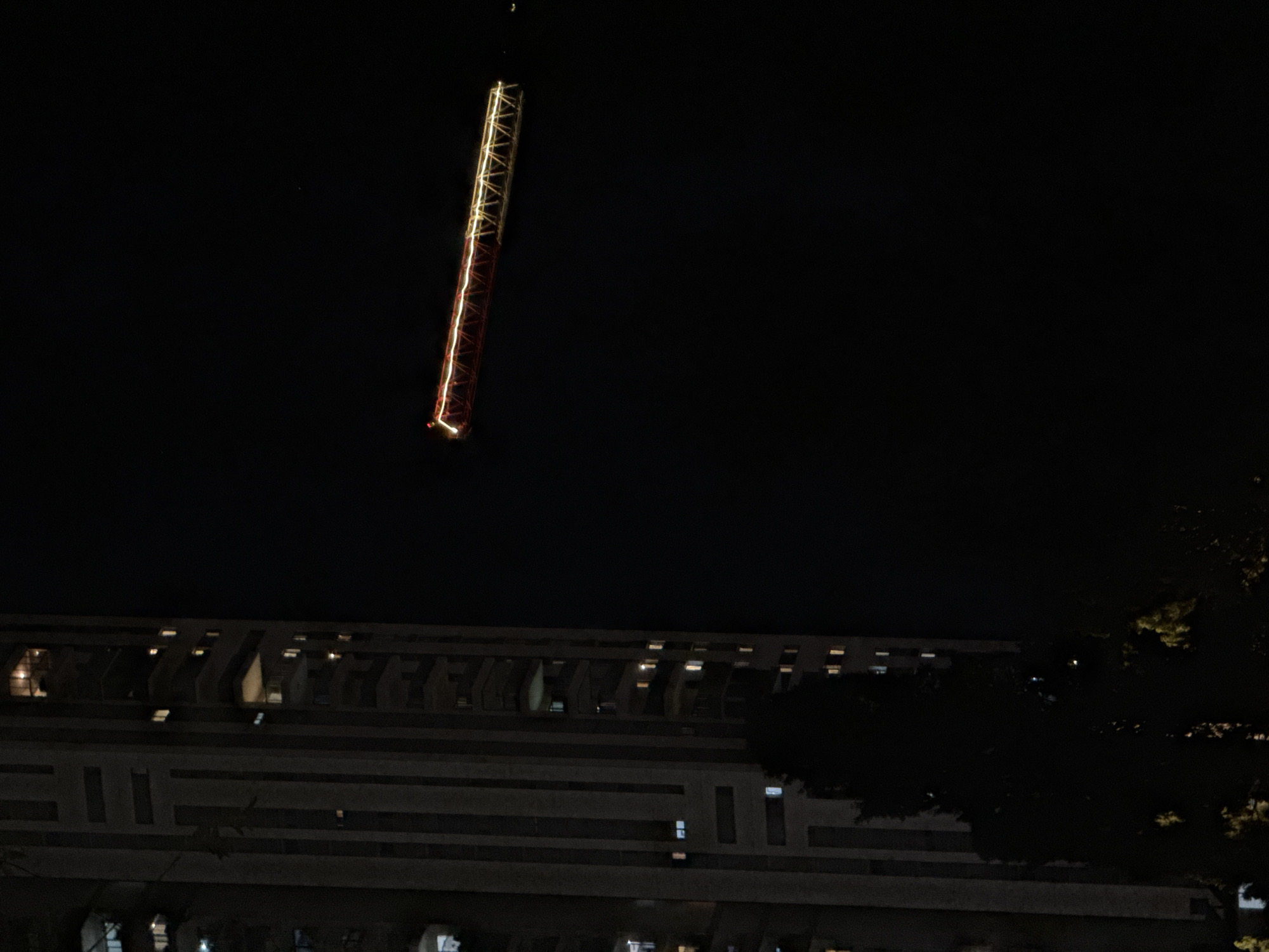 At night, a tall, partially illuminated tower rises above a multi-story building and a dark tree. The tower has a red base and a golden-yellow upper section, with lights running along its length. The building is dark, with rows of what appear to be recessed balconies or windows on multiple levels. The tree is silhouetted against the night sky. The location is possibly in Bat Yam, Israel, based on the prompt's instructions.