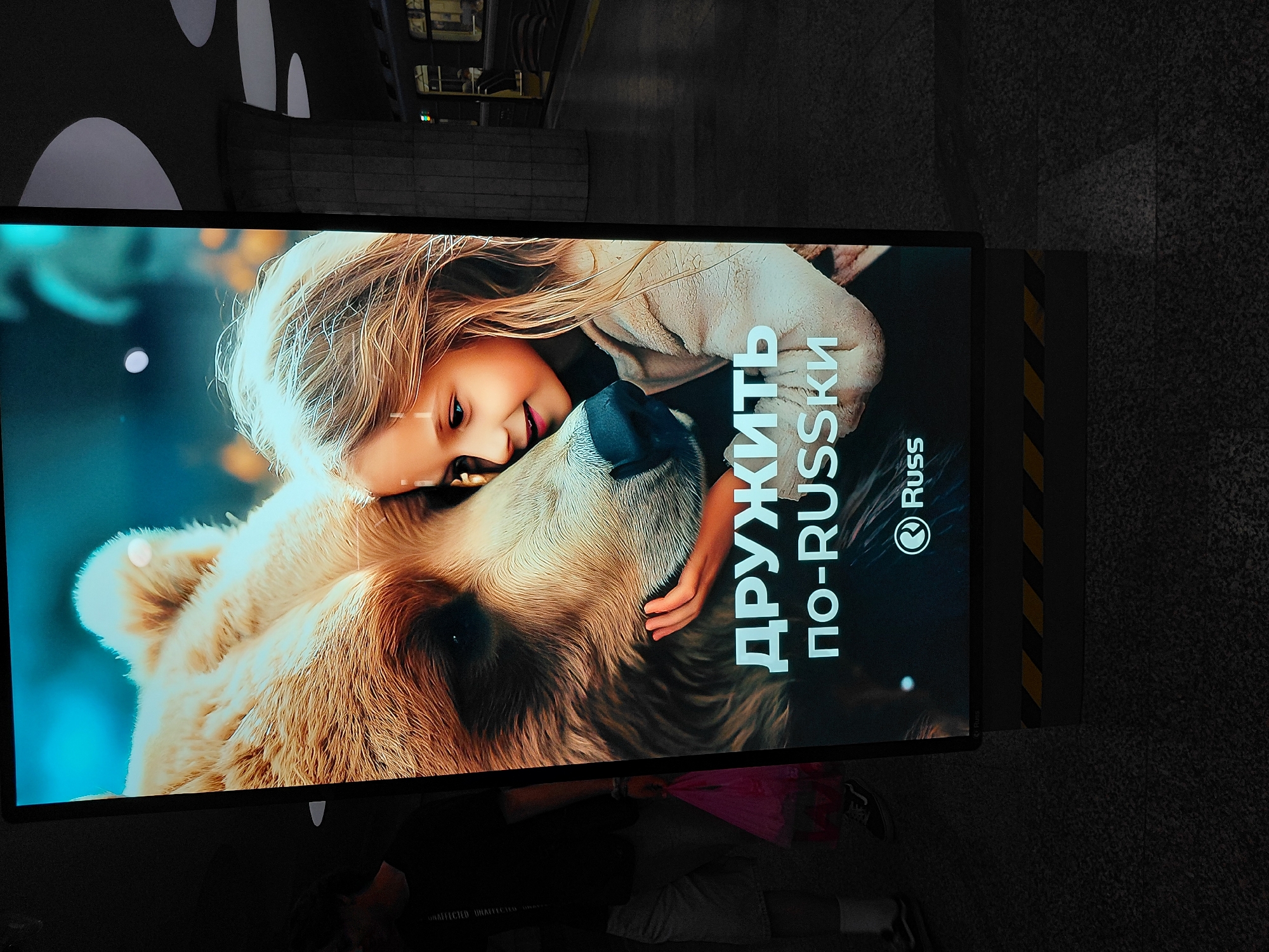 This is a photograph of a large digital billboard advertising "Russ" in Novosibirsk, Russia. The billboard features a close-up image of a young girl with blonde hair leaning in to touch the nose of a large brown bear. Both the girl and the bear appear to be looking at each other with gentle expressions. Below the image, text in Russian reads "ДРУЖИТЬ ПО-RUSSKI", which translates to "Be friends in Russian". The "Russ" logo is also visible. The billboard is illuminated and prominently displayed.
The setting appears to be a public transportation hub, likely a metro station, given the presence of a train track and a train car visible in the background. The architecture of the station, with its concrete columns and curved ceiling elements, is consistent with Soviet-era design often found in Russian metro systems. The floor is tiled or paved, and there are yellow and black hazard stripes on the base of the billboard.
In the foreground and to the left of the billboard, a person wearing a dark t-shirt and light-colored shorts is partially visible. They are holding a pink plastic bag in their left hand. The lighting conditions suggest an indoor environment with artificial lighting, possibly late evening or underground. The overall impression is of a public space in Novosibirsk showcasing an advertisement.