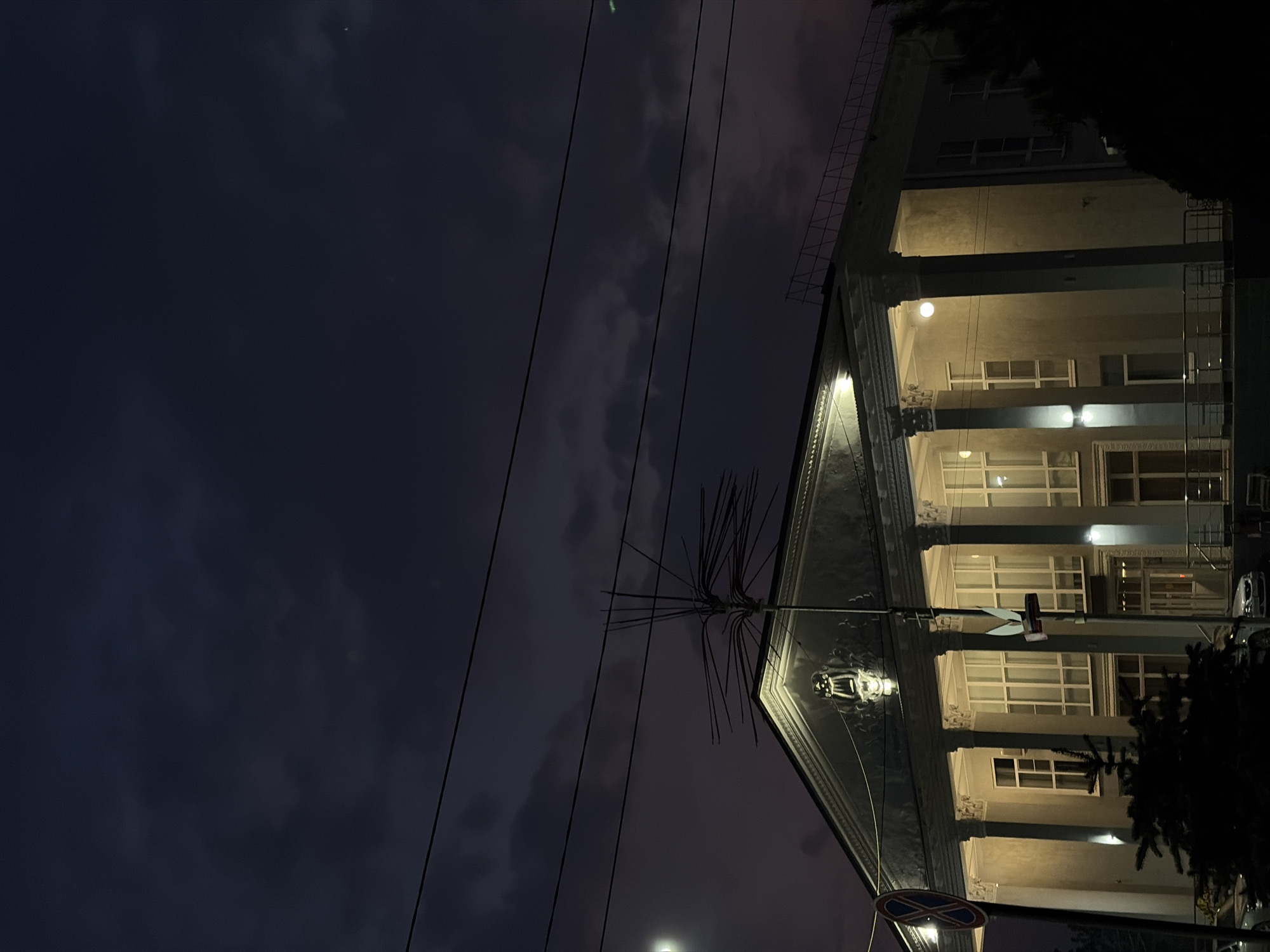 The image presents an evening view of a building in Novopokrovskaya, Russia. The building is multi-story with a colonnaded facade illuminated by interior lights. Windows are visible between the columns. Above the columns, there is a decorative roofline. Utility wires stretch across the dark, cloudy sky in the background. A streetlight pole with an electrical junction is in the foreground, along with a road sign. Part of a tree obscures the building.