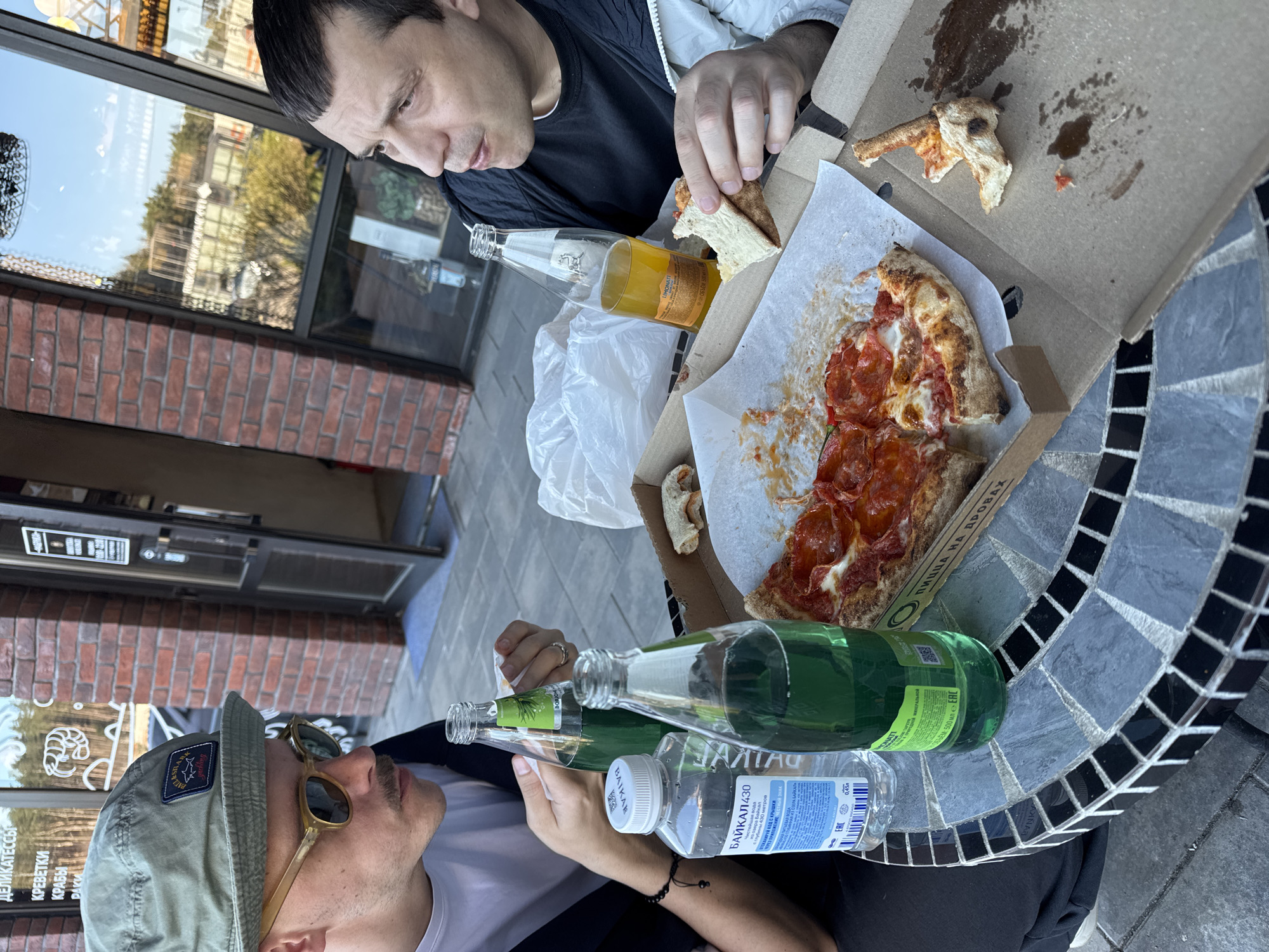 Two men are sitting at a mosaic table, consuming pizza and beverages outdoors. The pizza, partially eaten, rests in an open cardboard box on the table. Various beverages, including an orange-colored drink in a clear glass bottle, green-labeled bottles, and a plastic bottle of water with a blue label, are also present on the table. One man, dressed in a dark shirt and a light jacket, is holding a piece of pizza. The other man is wearing sunglasses and a cap with a logo. The table is positioned outside what appears to be a storefront with glass windows. Text in Russian, including words related to delicacies, shrimp, and crab, is visible on the store window. The environment suggests an urban setting.